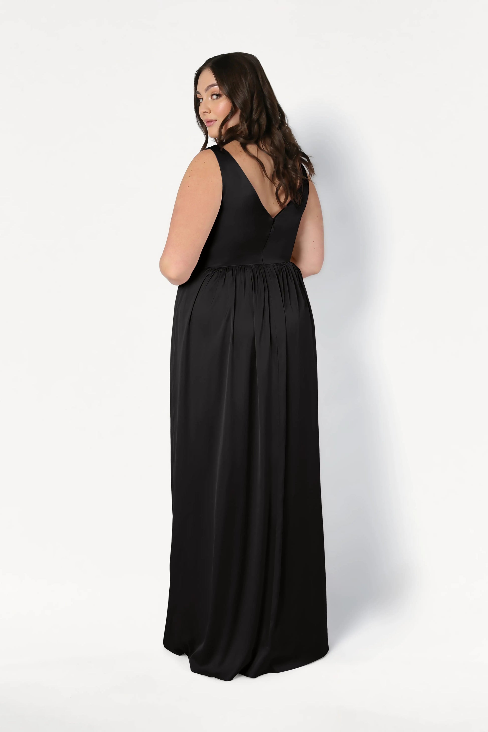 A woman models the TH&TH Carmen Bridesmaid Dress in Black Satin, a sleeveless floor-length gown, standing against a plain white background and glancing over her shoulder with a slight smile.