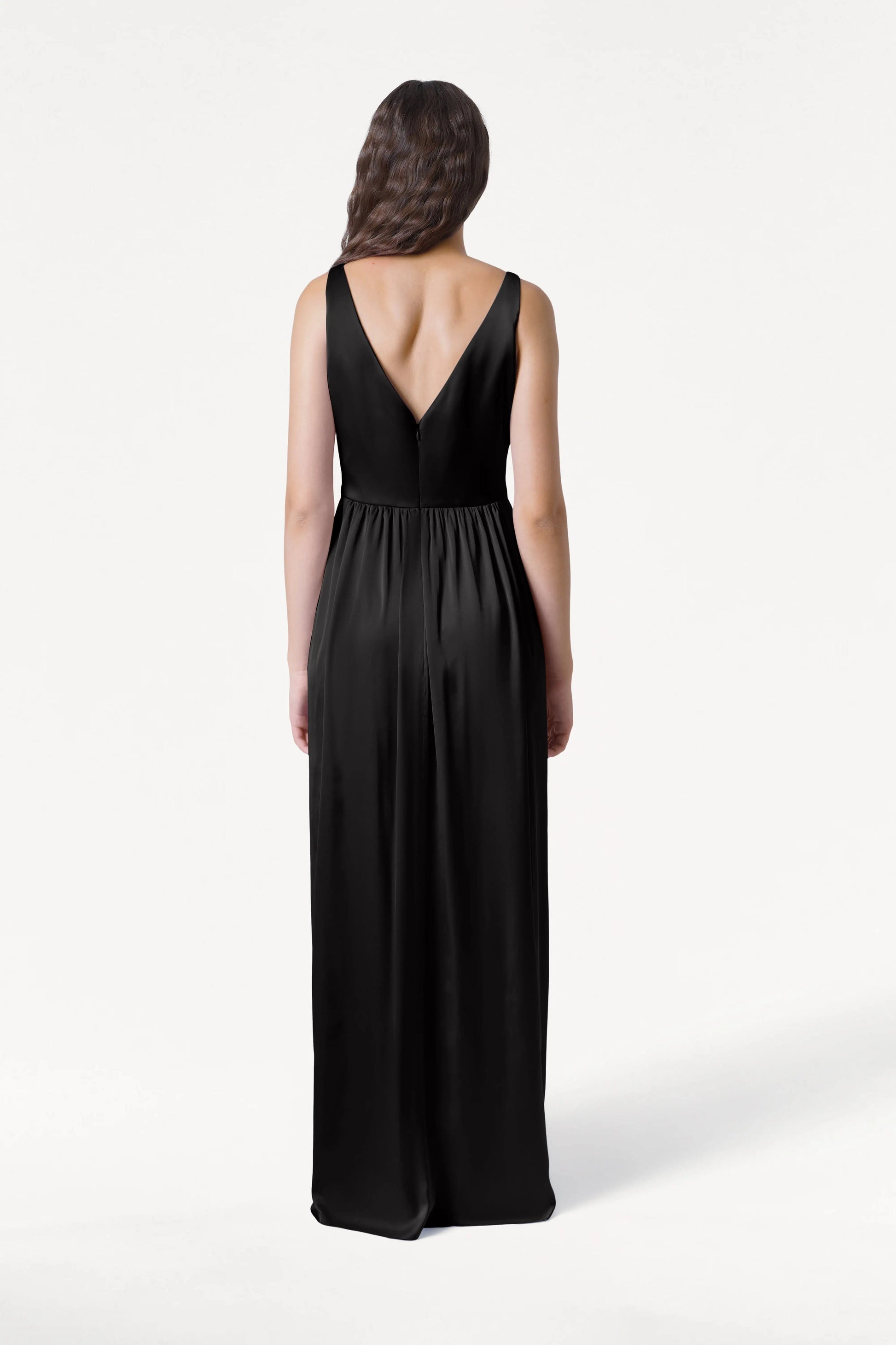 A woman with long, wavy brown hair faces away, modeling the TH&TH Carmen Bridesmaid Dress in Black Satin. The gown features a deep V-back and is ideal for a modern bridal party, shown against a plain white background.