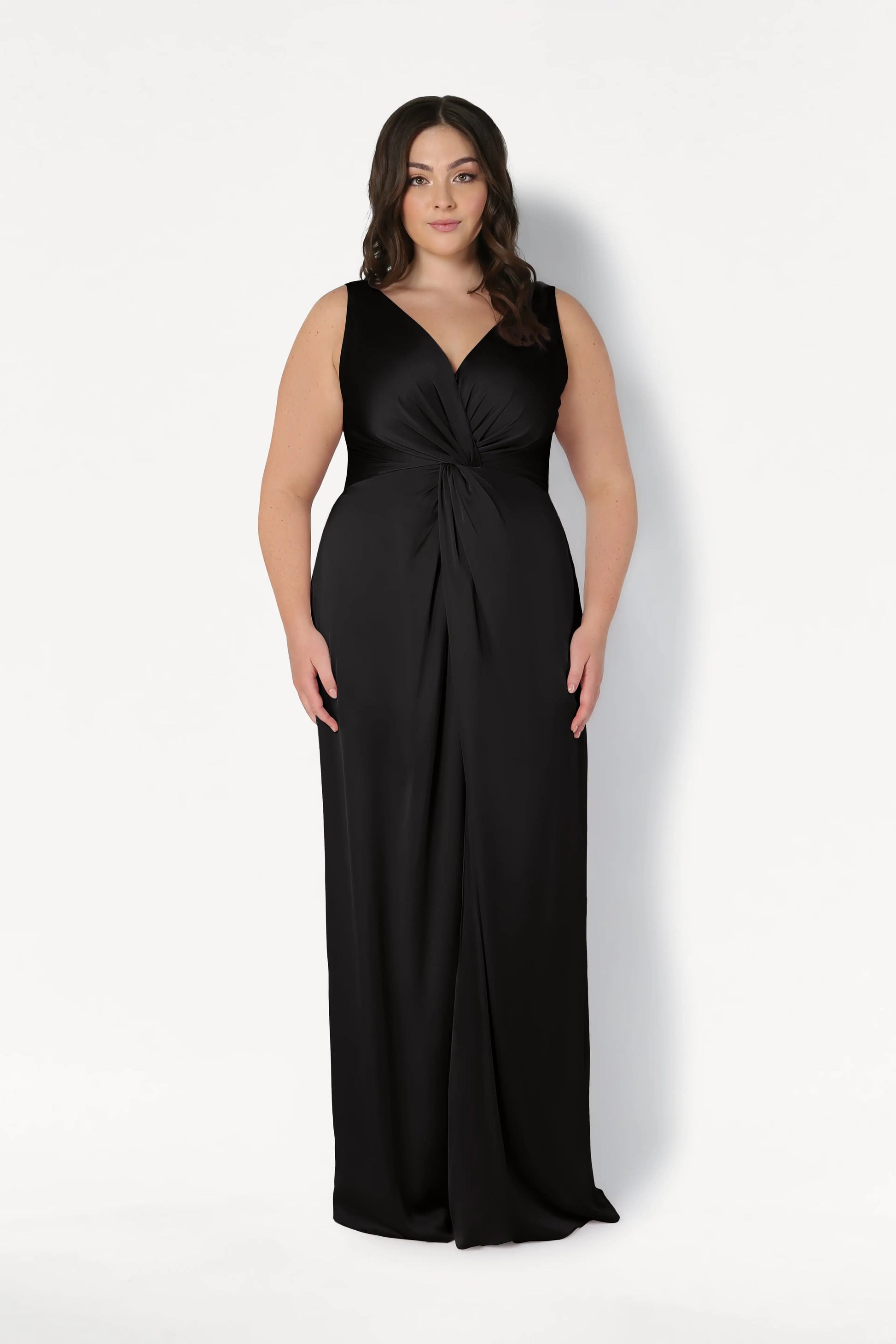 A woman with long dark hair models the TH&TH Carmen Bridesmaid Dress in Black Satin, a sleeveless, floor-length gown with a twist-knot front and V-neckline, standing against a plain white background.