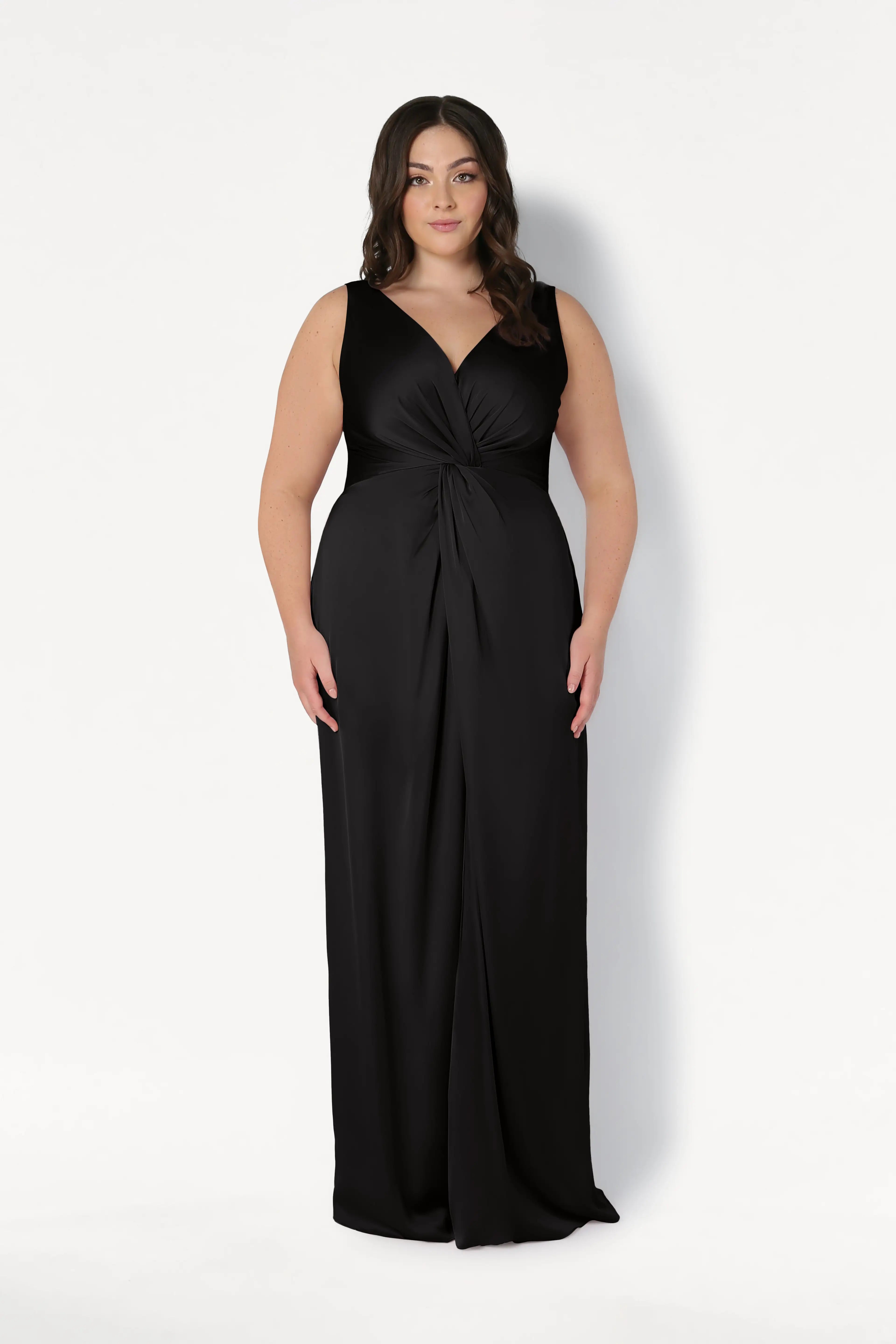 A woman with long dark hair models the TH&TH Carmen Bridesmaid Dress in Black Satin, a sleeveless, floor-length gown with a twist-knot front and V-neckline, standing against a plain white background.