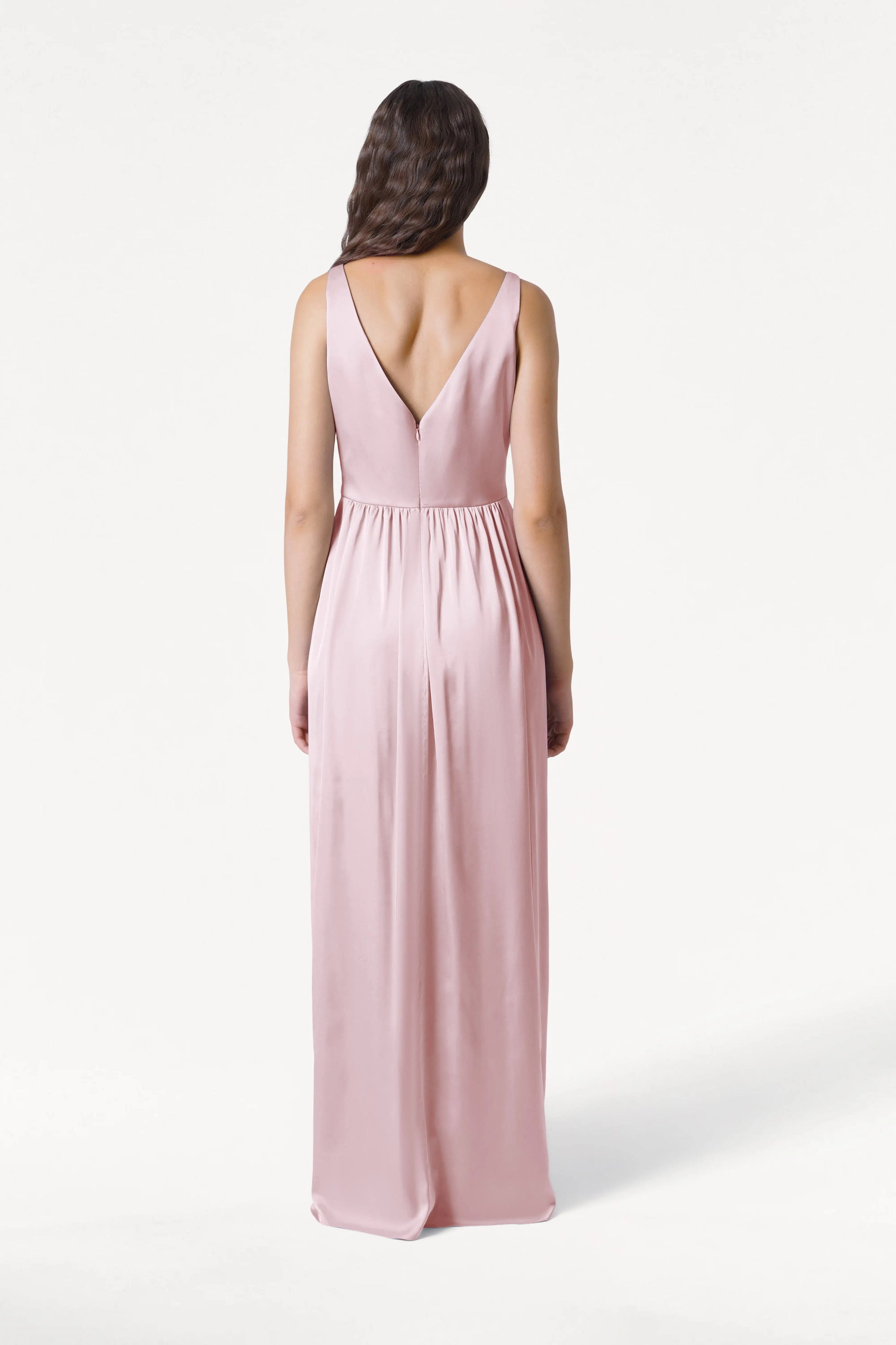 A woman faces away from the camera, wearing the Carmen Bridesmaid Dress in Blush Pink Satin by TH&TH—a long, sleeveless gown with a deep V-back and gathered waist—her loose waves complementing the modern bridal look against a white background.