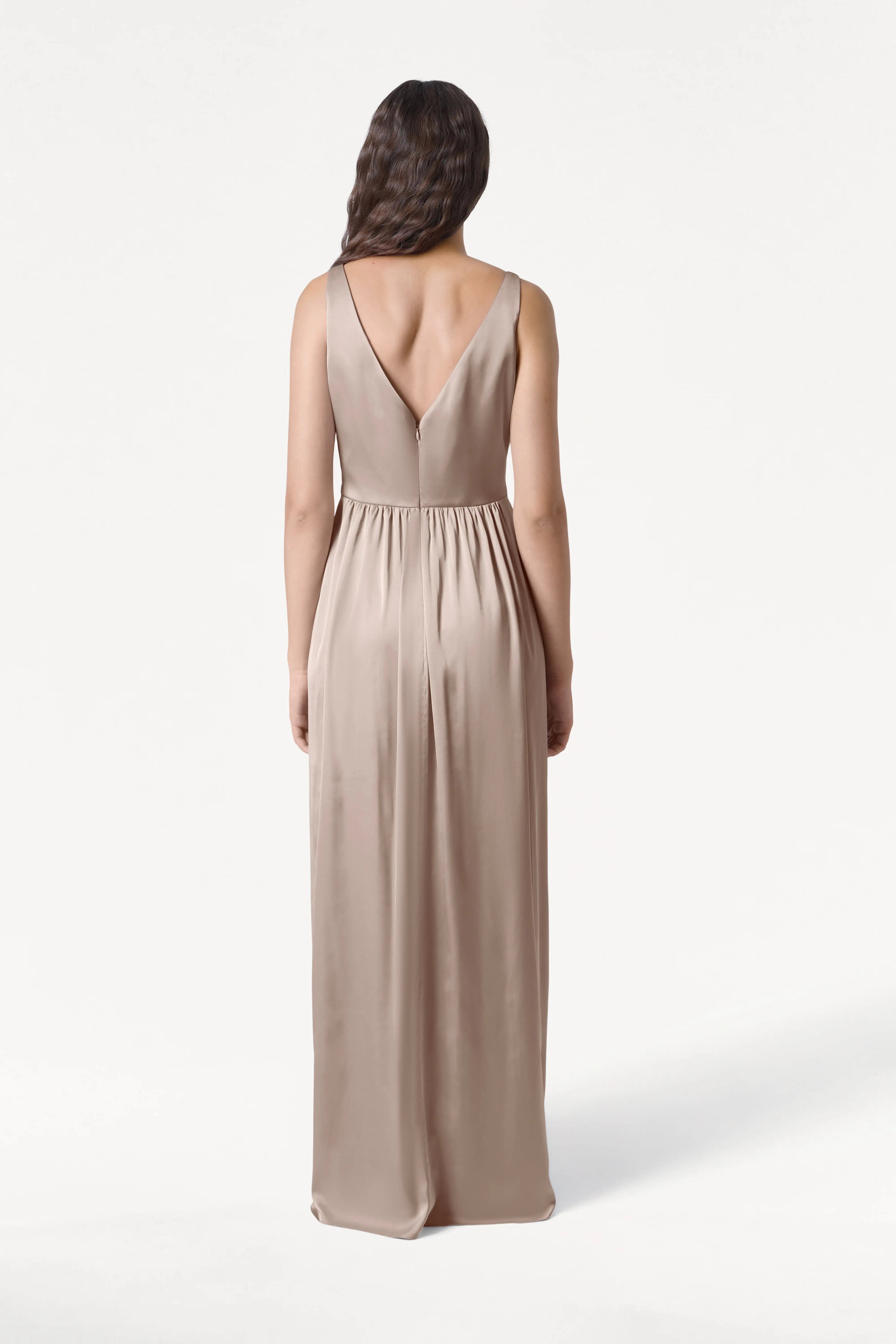 A woman with long, wavy hair stands facing away in the TH&TH Carmen Bridesmaid Dress—Champagne Sky Satin, a sleeveless, floor-length gown with a deep V-back and gathered waist, ideal for modern bridal parties against a white backdrop.