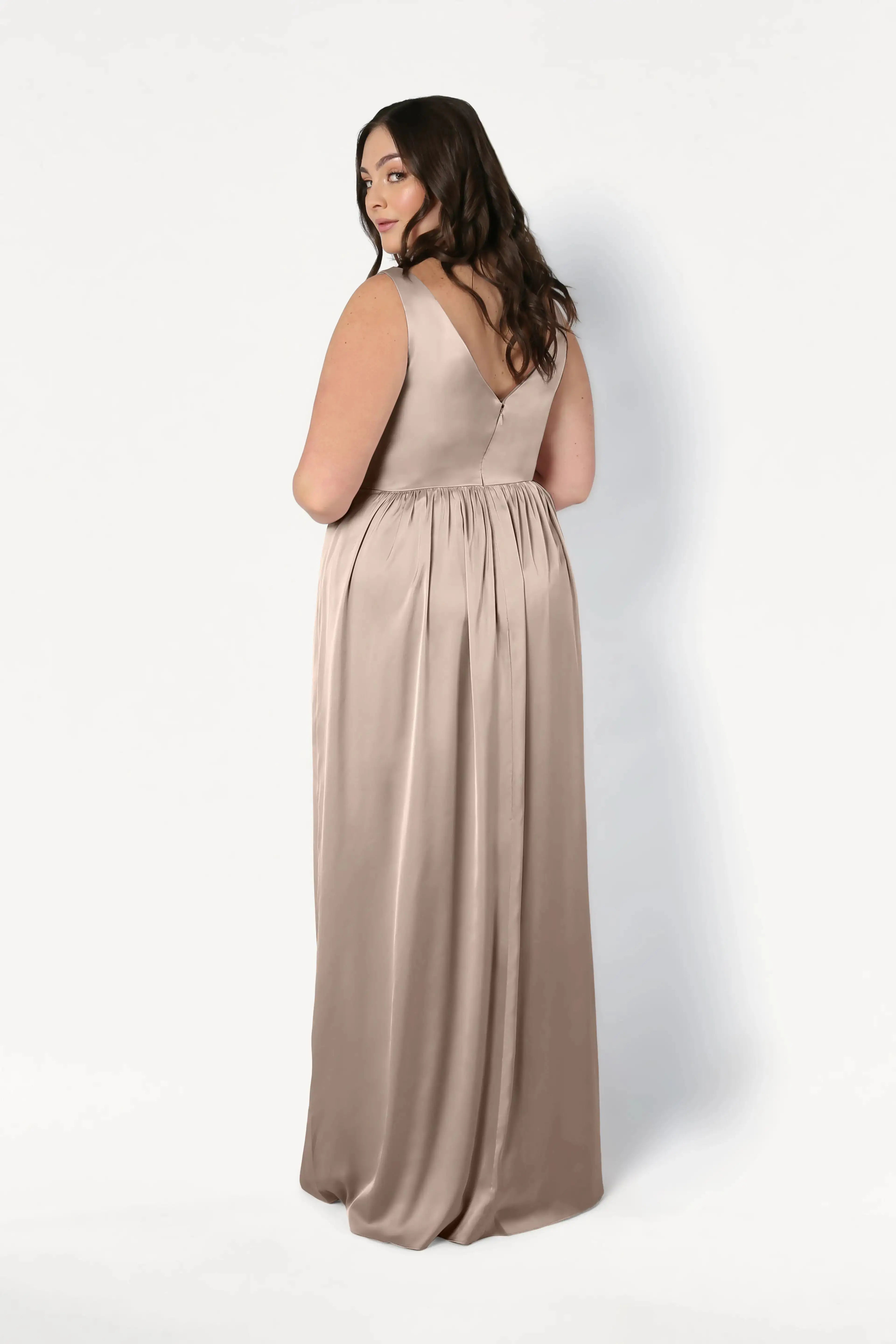 A woman with long, wavy brown hair stands slightly turned away, wearing the TH&TH Carmen Bridesmaid Dress in Champagne Sky Satin—a stylish V-back gown ideal for modern bridal parties—against a plain white background.