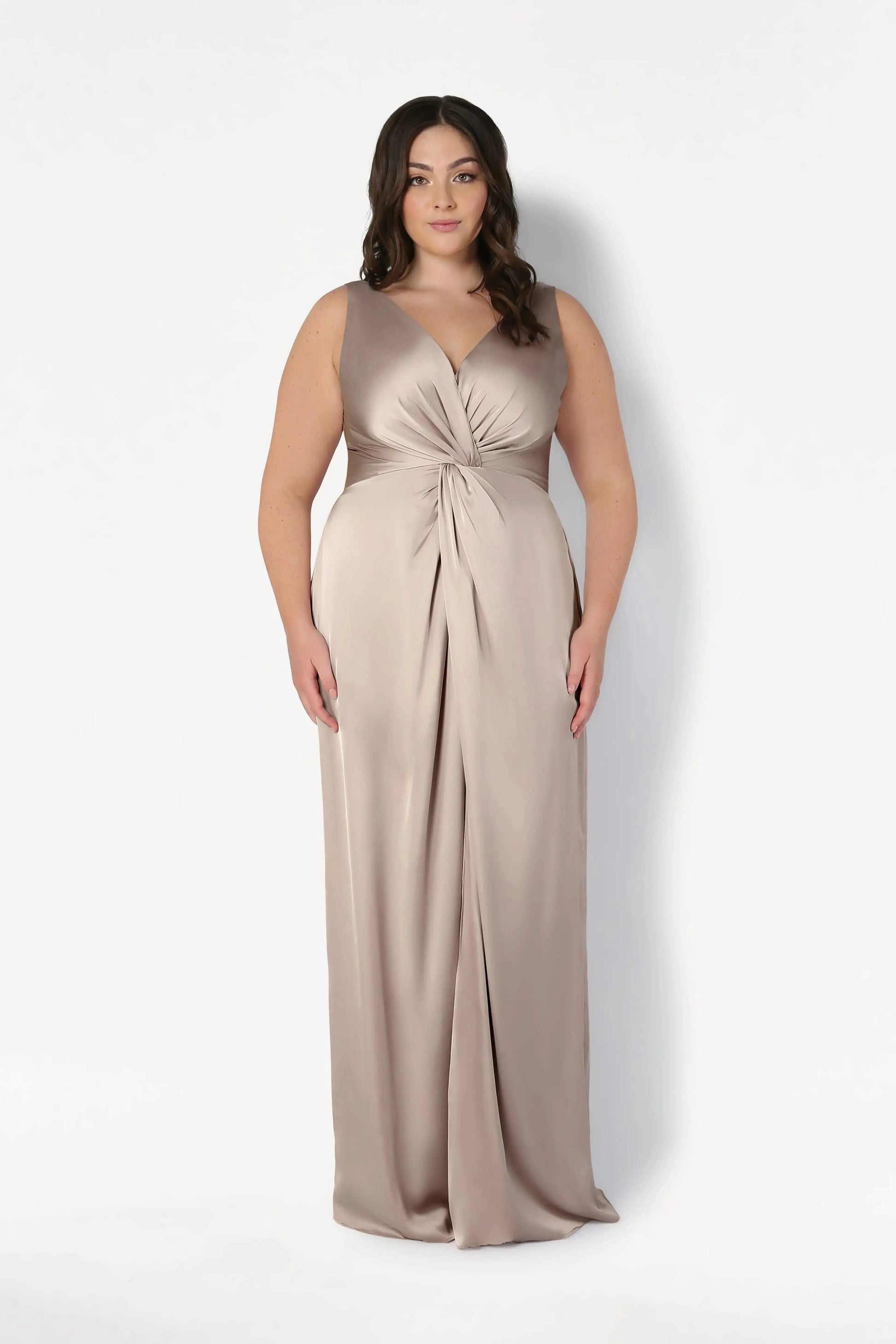 A woman with long, wavy dark hair wears the TH&TH Carmen Bridesmaid Dress in Champagne Sky Satin, a sleeveless, floor-length gown with a gathered knot waist, standing against a white background and smiling—a chic choice for modern bridal parties.