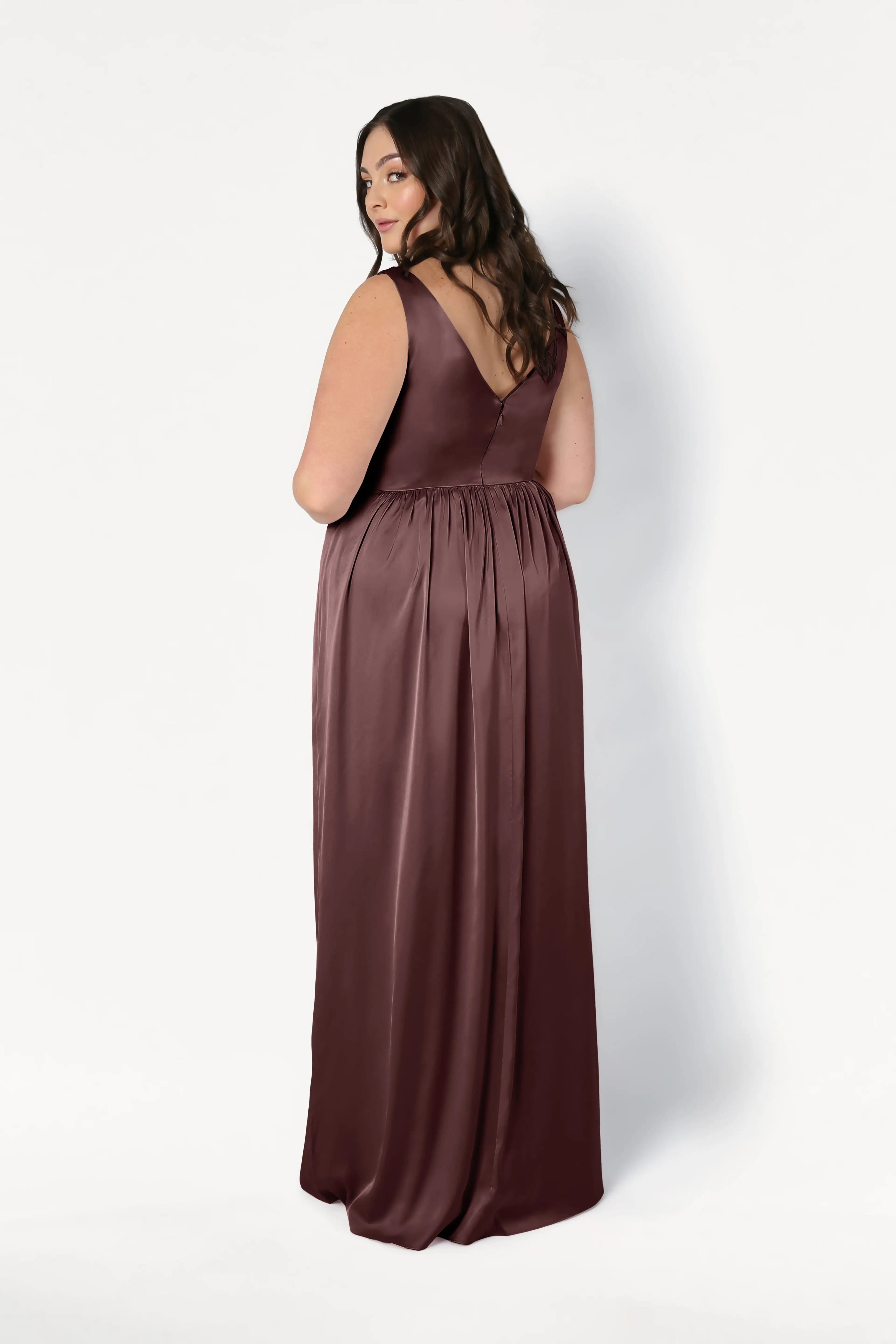 A woman with long brown hair wears the Carmen Bridesmaid Dress in chocolate brown satin by TH&TH, featuring a sleeveless, floor-length design and V-shaped back—ideal for modern bridal parties against a plain white background.