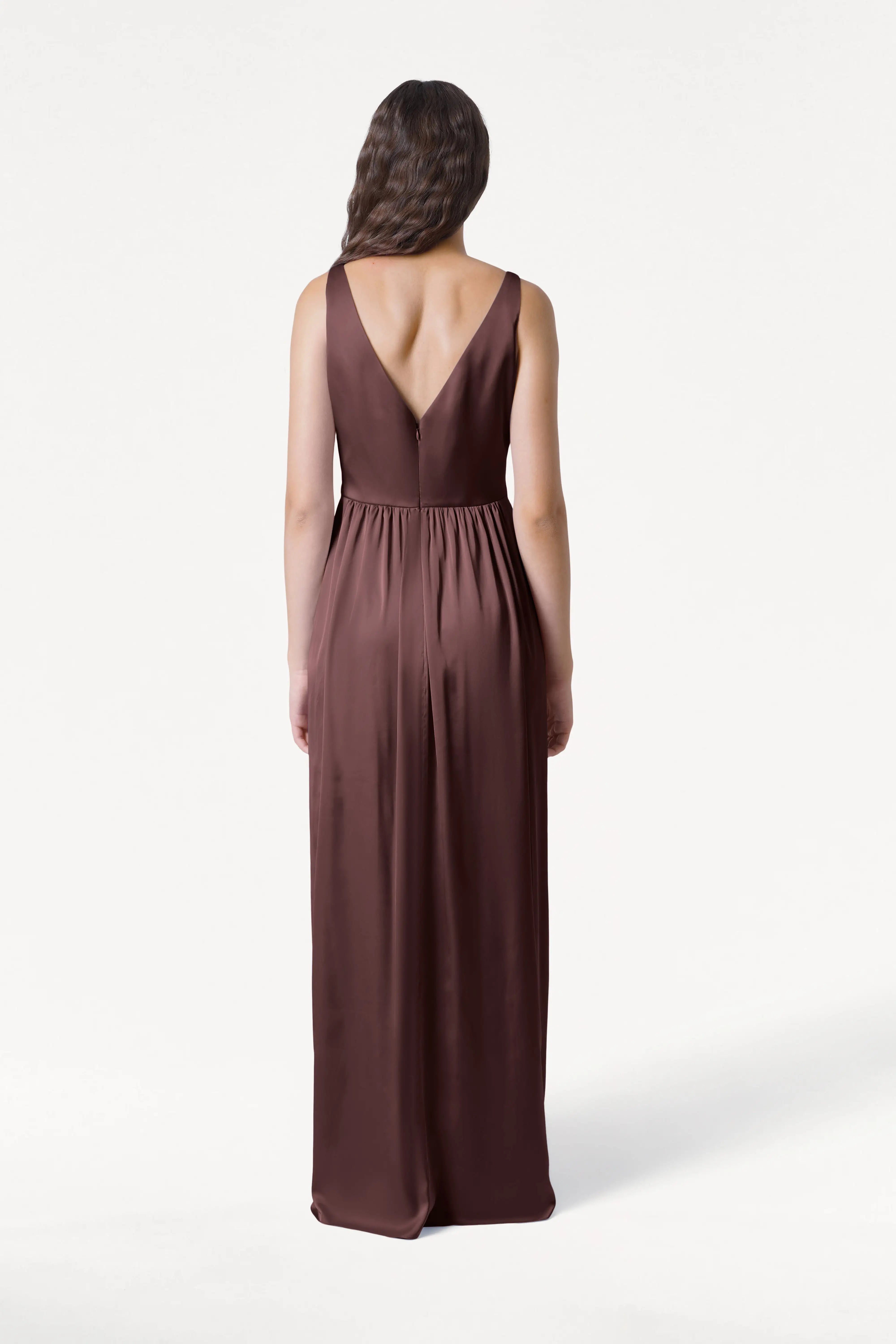 A woman with long wavy brown hair stands with her back to the camera, wearing the TH&TH Carmen Bridesmaid Dress in Chocolate Brown Satin—a sleeveless, floor-length gown with a V-shaped back, perfect for modern bridal parties.