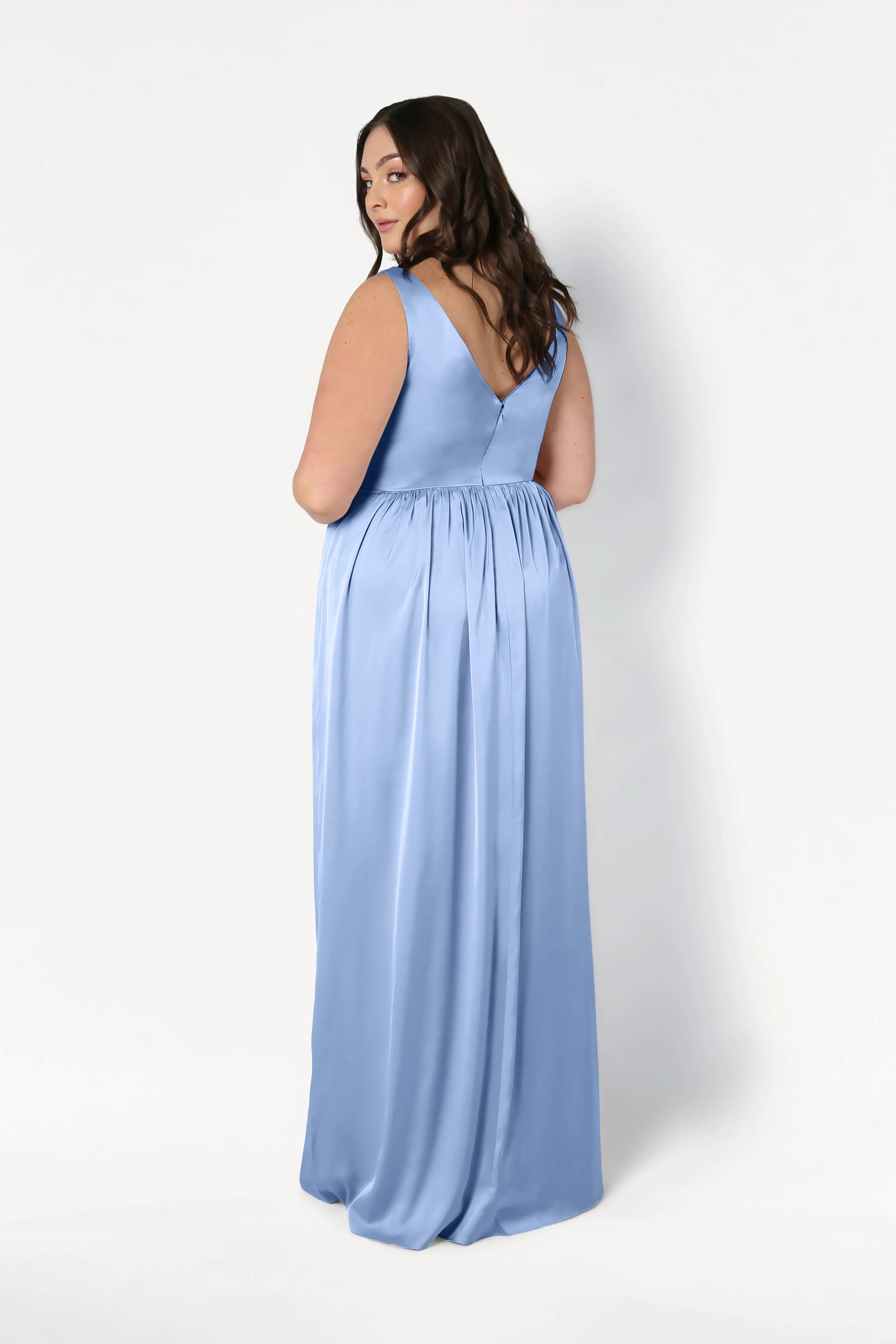A plus-size model wears the Carmen Bridesmaid Dress in Dusty Blue Satin by TH&TH, featuring a V-back, gathered waist, and flowing skirt. The floor-length gown is shown from behind against a white background—ideal for modern bridal parties.