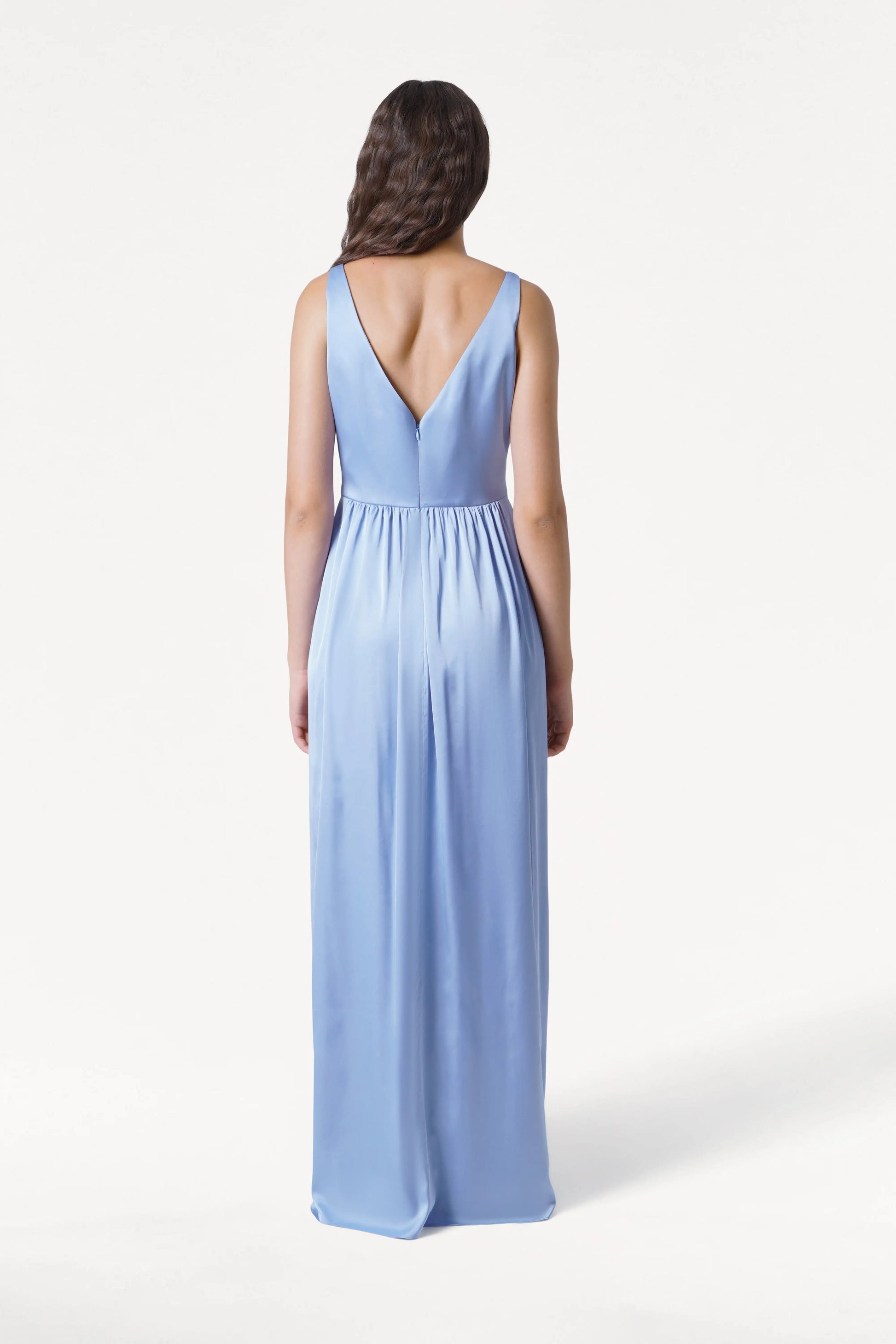 A woman with long wavy hair, seen from the back, wears the TH&TH Carmen Bridesmaid Dress in Dusty Blue satin—a sleeveless, floor-length gown with a deep V-back—posed against a plain white background.