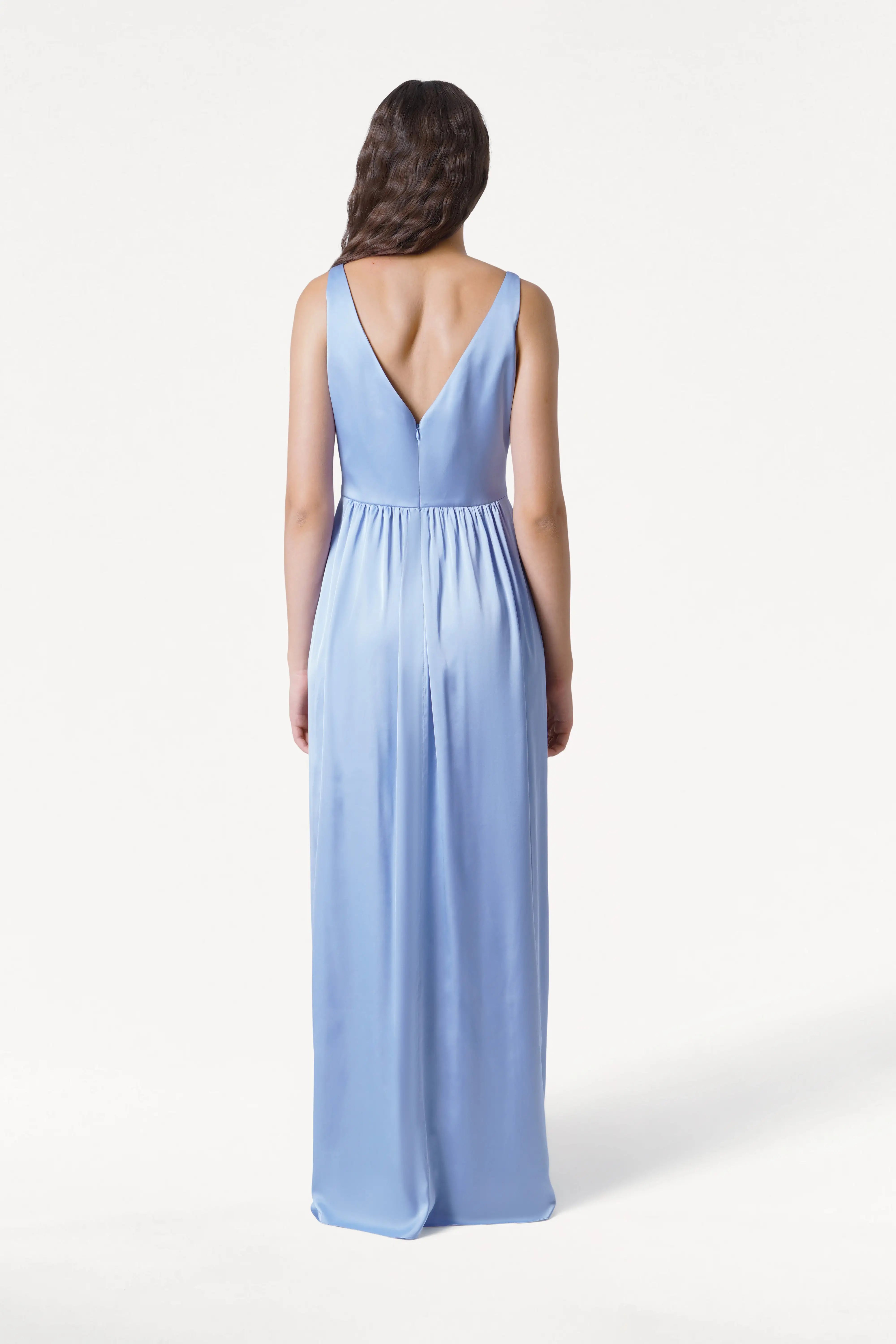 A woman with long wavy hair, seen from the back, wears the TH&TH Carmen Bridesmaid Dress in Dusty Blue satin—a sleeveless, floor-length gown with a deep V-back—posed against a plain white background.