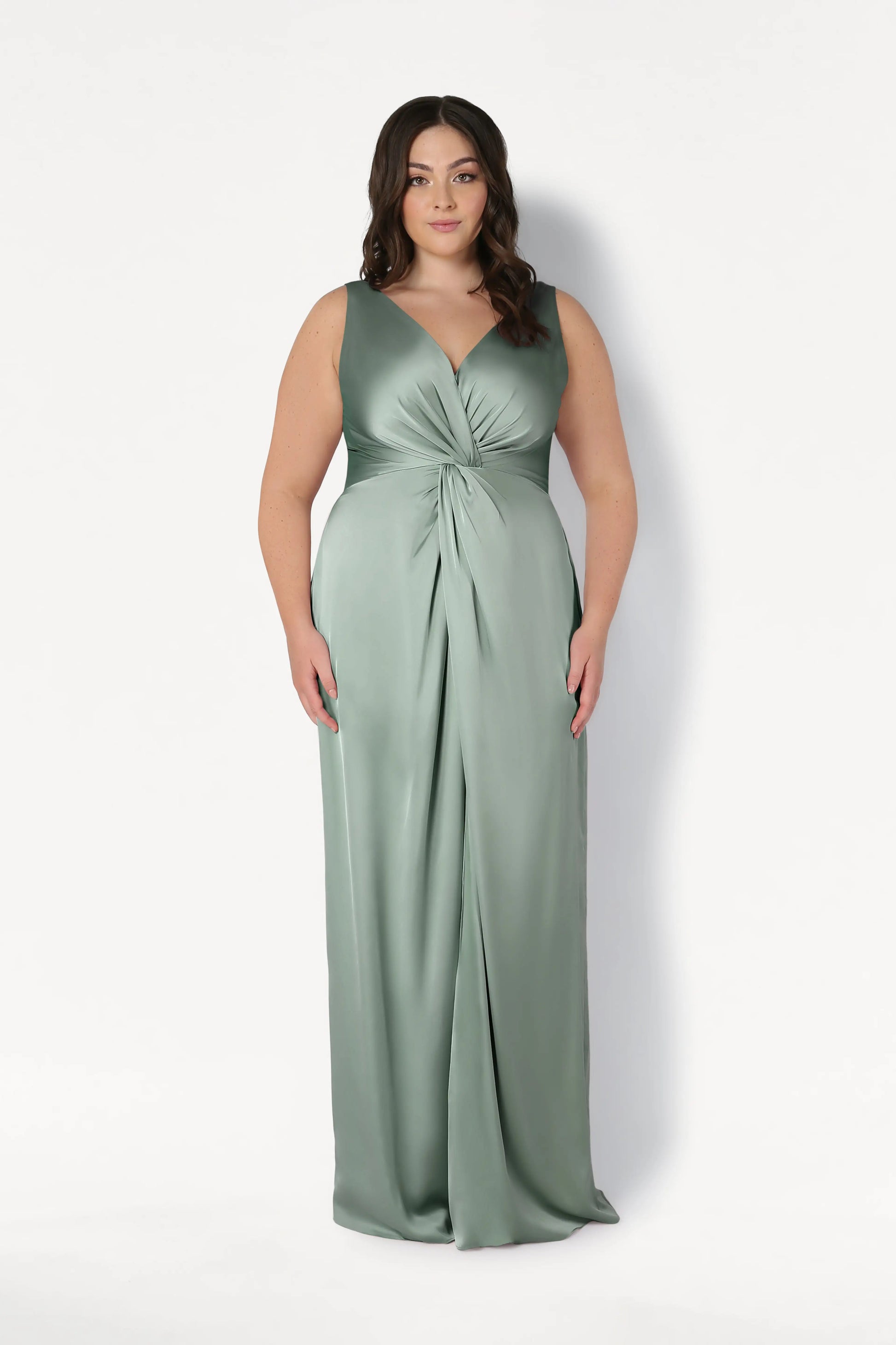 A woman wears the Carmen Bridesmaid Dress in Dusty Eucalyptus Satin by TH&TH, a sleeveless, floor-length gown with a v-neckline and gathered knot waist, shown against a white background—ideal for modern bridal parties.