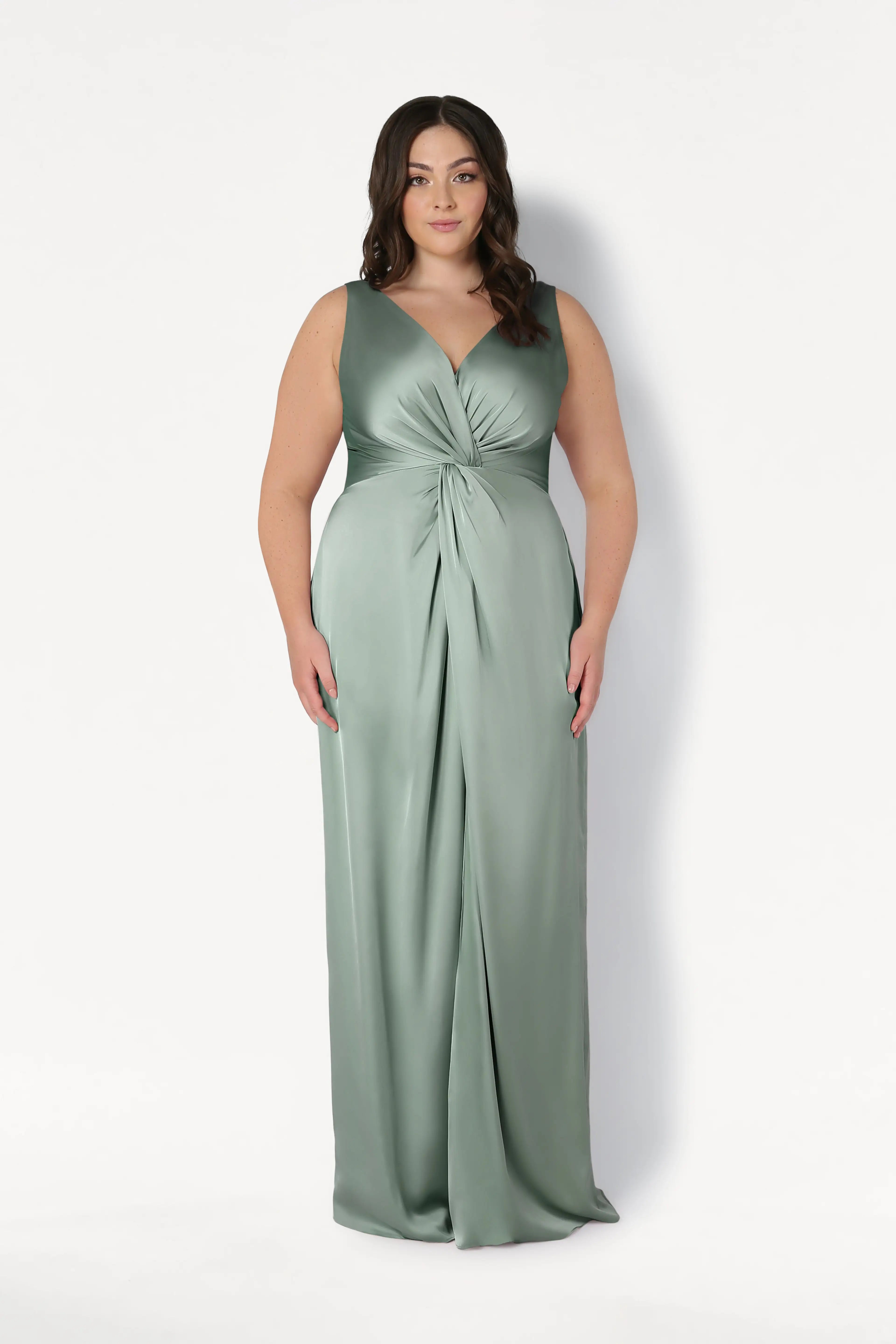 A woman wears the Carmen Bridesmaid Dress in Dusty Eucalyptus Satin by TH&TH, a sleeveless, floor-length gown with a v-neckline and gathered knot waist, shown against a white background—ideal for modern bridal parties.