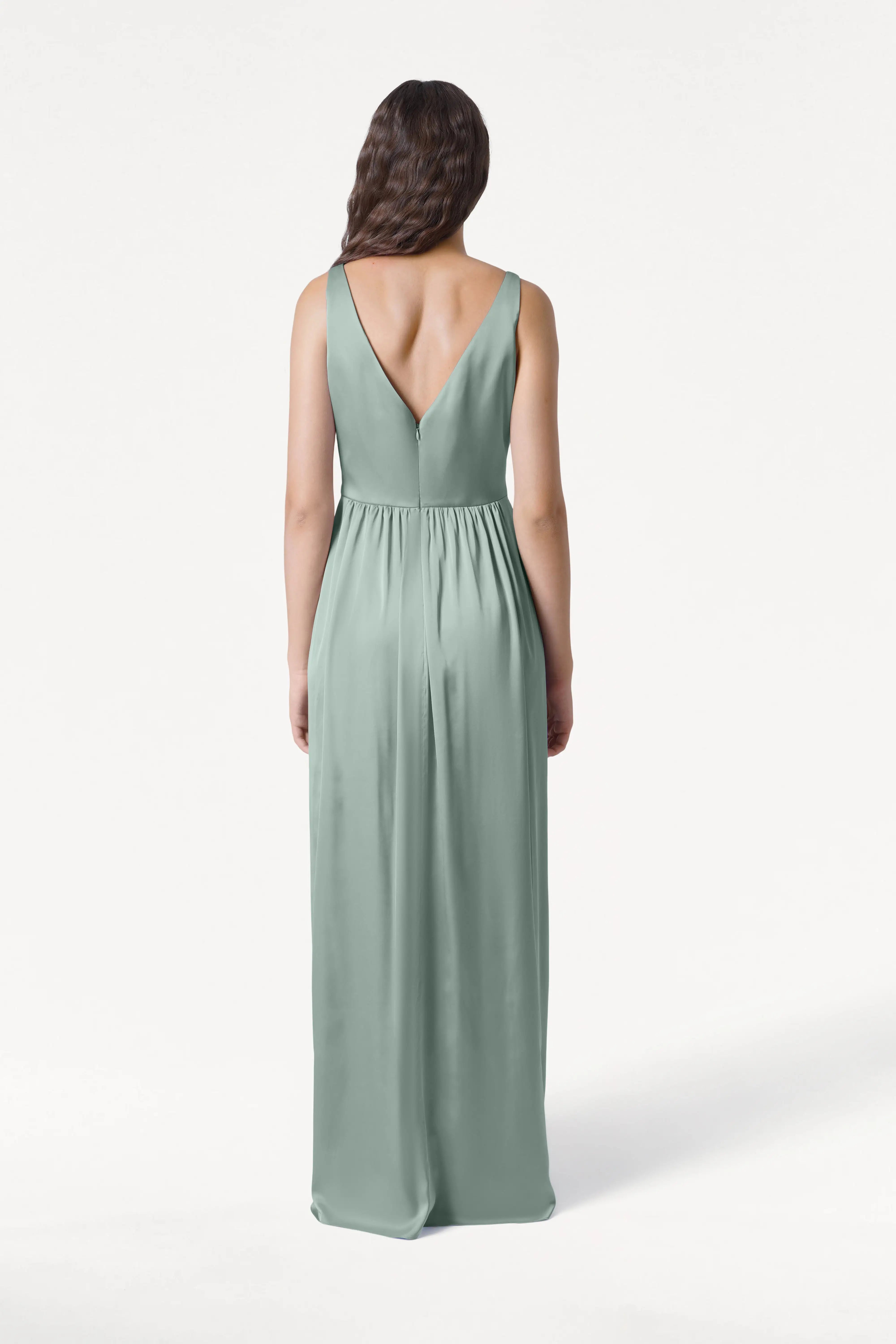 A woman with long, wavy brown hair stands facing away in the TH&TH Carmen Bridesmaid Dress— a sleeveless, floor-length dusty eucalyptus satin gown with a deep V-back. The background is plain white, making this a chic option for modern bridal parties.