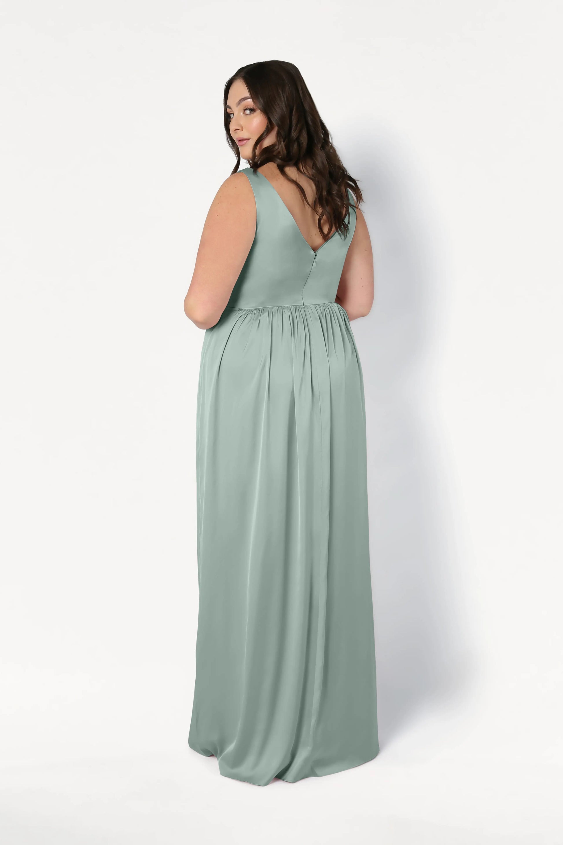 A woman with long dark hair models the TH&TH Carmen Bridesmaid Dress in Dusty Eucalyptus Satin, a sleeveless, floor-length gown with a V-shaped back—an elegant choice for contemporary bridal parties, shown against a plain white background.