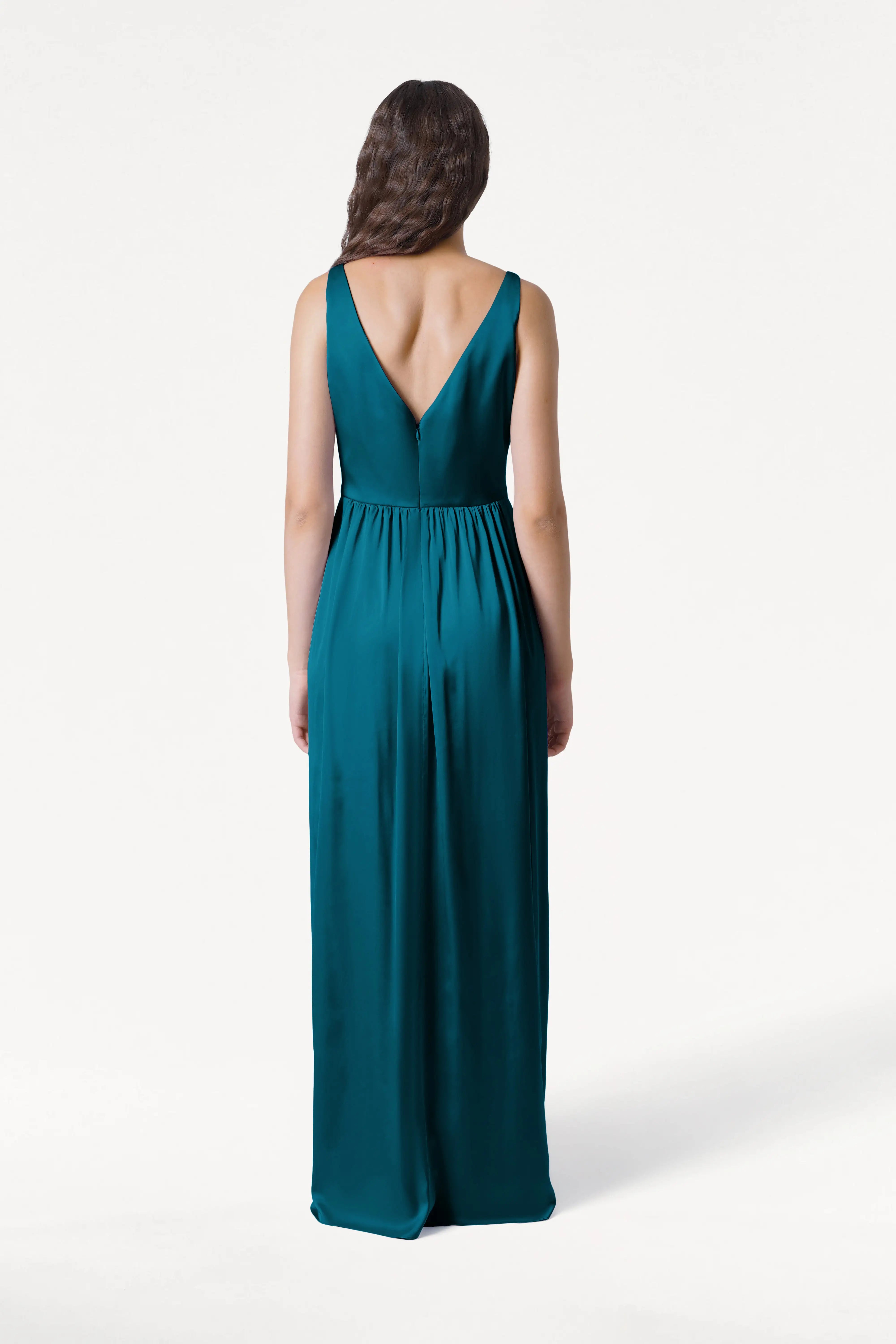 A woman with long wavy brown hair models the TH&TH Carmen Bridesmaid Dress in Emerald Dawn Satin, a sleeveless gown with a deep V-back, standing and facing away against a plain white background.