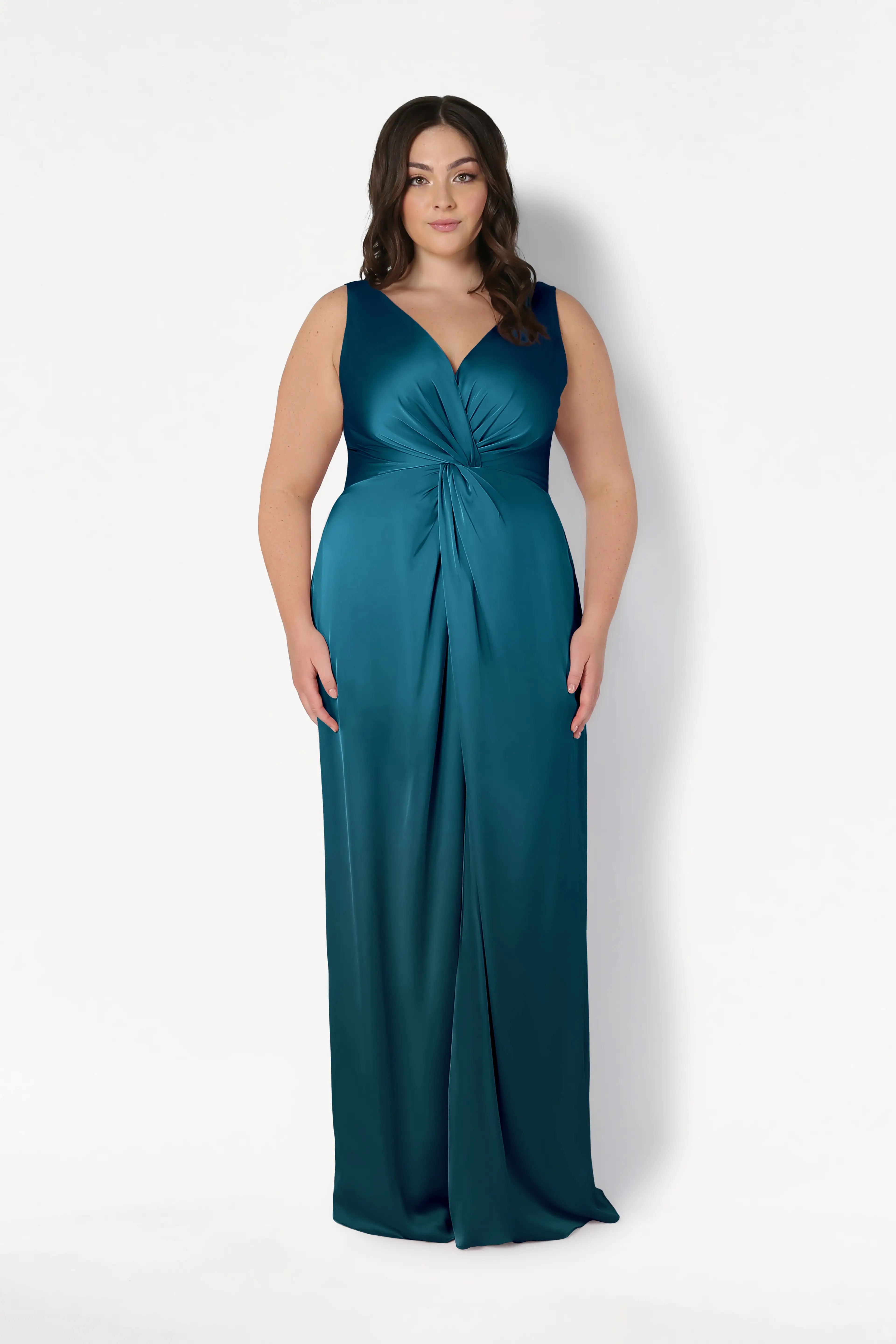 A woman with long dark hair wears the TH&TH Carmen Bridesmaid Dress in Emerald Dawn Satin, a sleeveless gown with a V-neckline and twist-knot waist detail, standing against a plain white background.