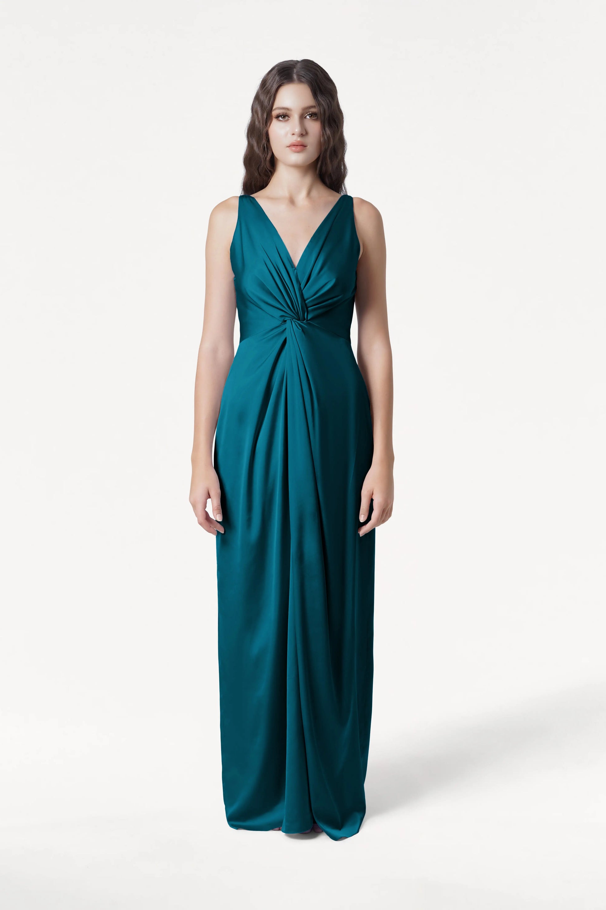 A woman with long, wavy brown hair and a neutral expression models the TH&TH Carmen Bridesmaid Dress in Emerald Dawn Satin—a sleeveless, floor-length gown with a deep V-neck and twist-knot front—against a plain white background.
