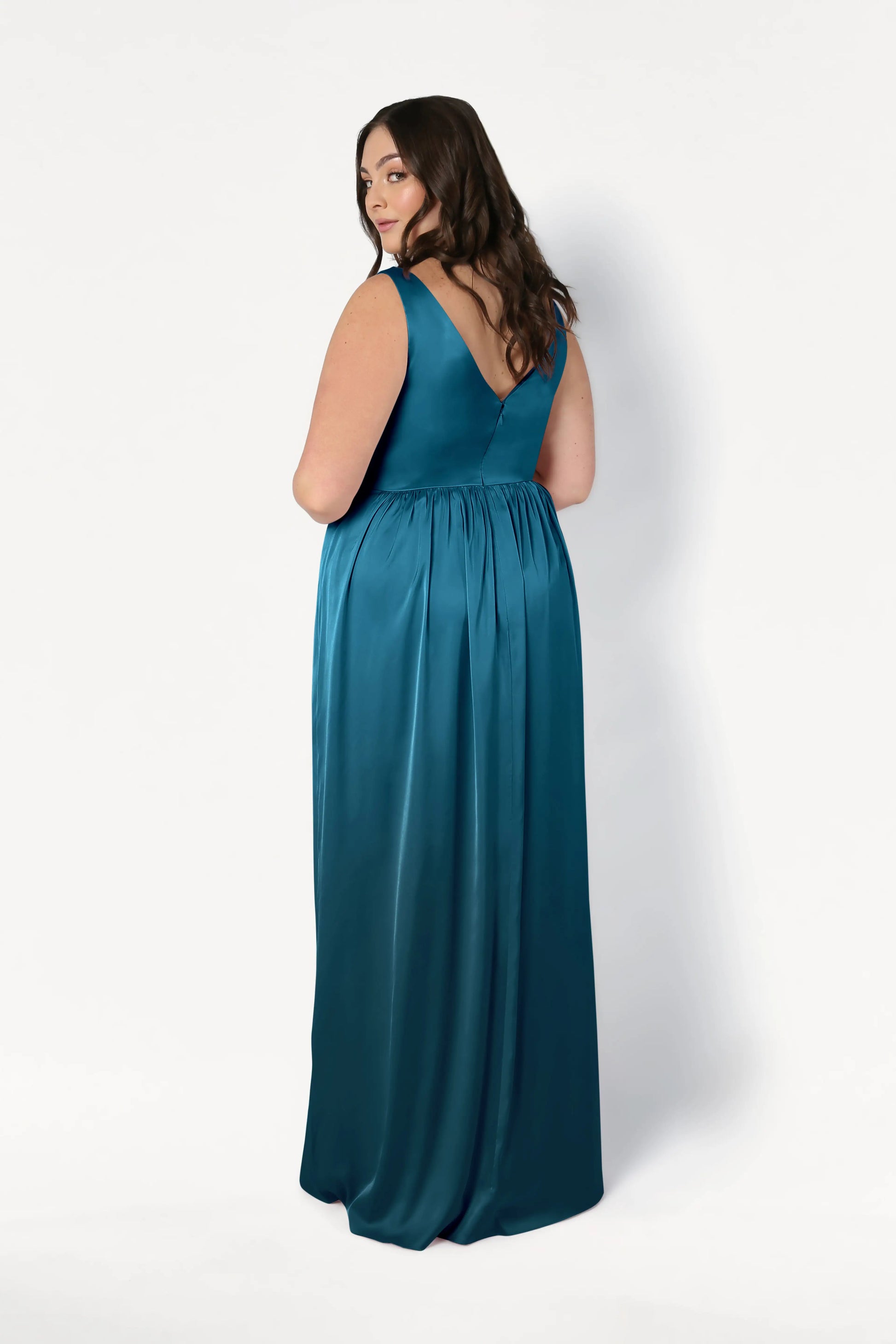 A woman with long brown hair models the TH&TH Carmen Bridesmaid Dress in Emerald Dawn Satin, showcasing its sleeveless, floor-length design and deep V back against a plain white background—ideal for modern bridal parties.
