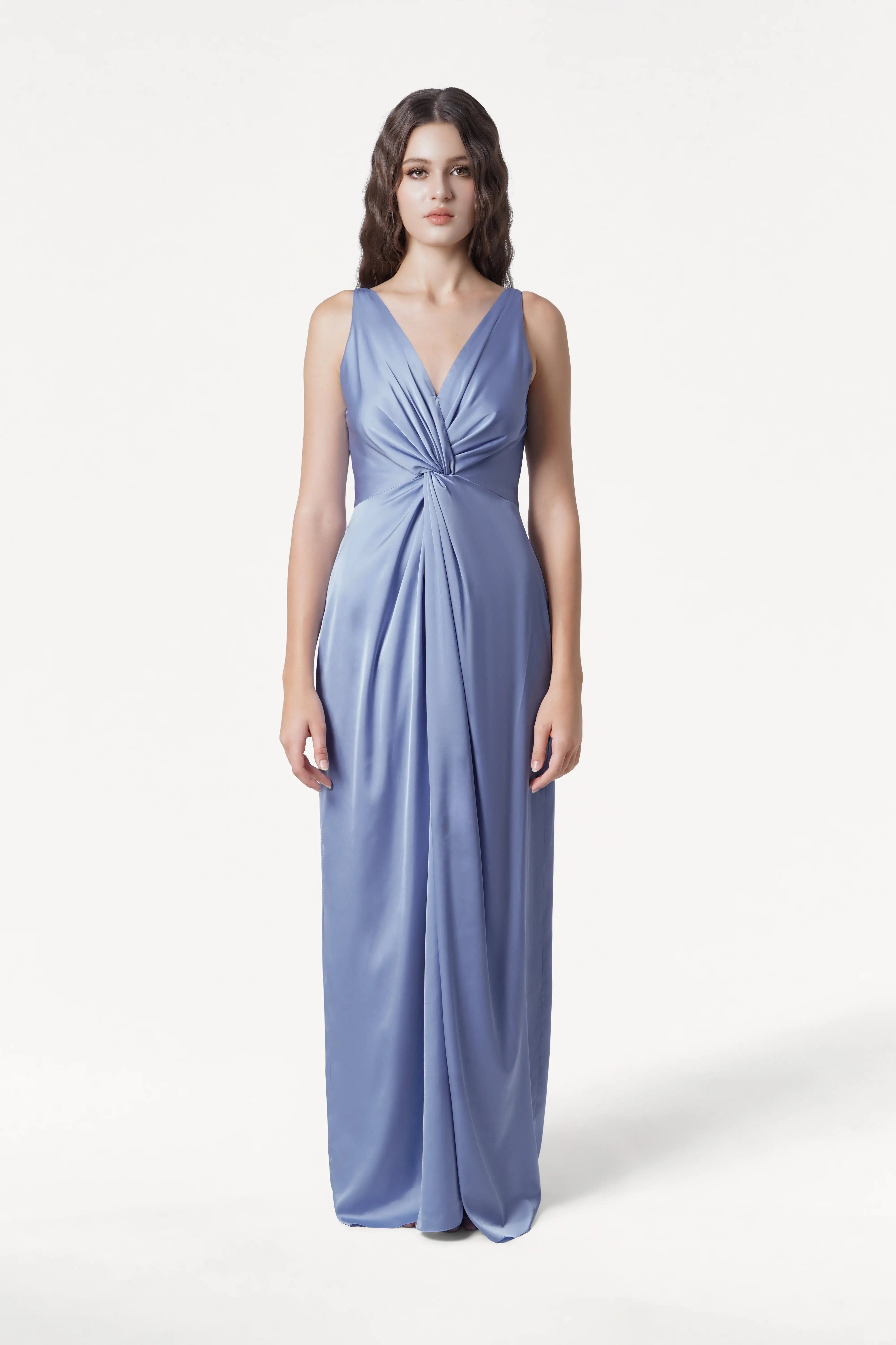 A woman poses against a plain white background in the Carmen Bridesmaid Dress by TH&TH—a sleeveless, floor-length French Blue satin gown with a deep V-neck and an elegant twist-knot detail at the waist.