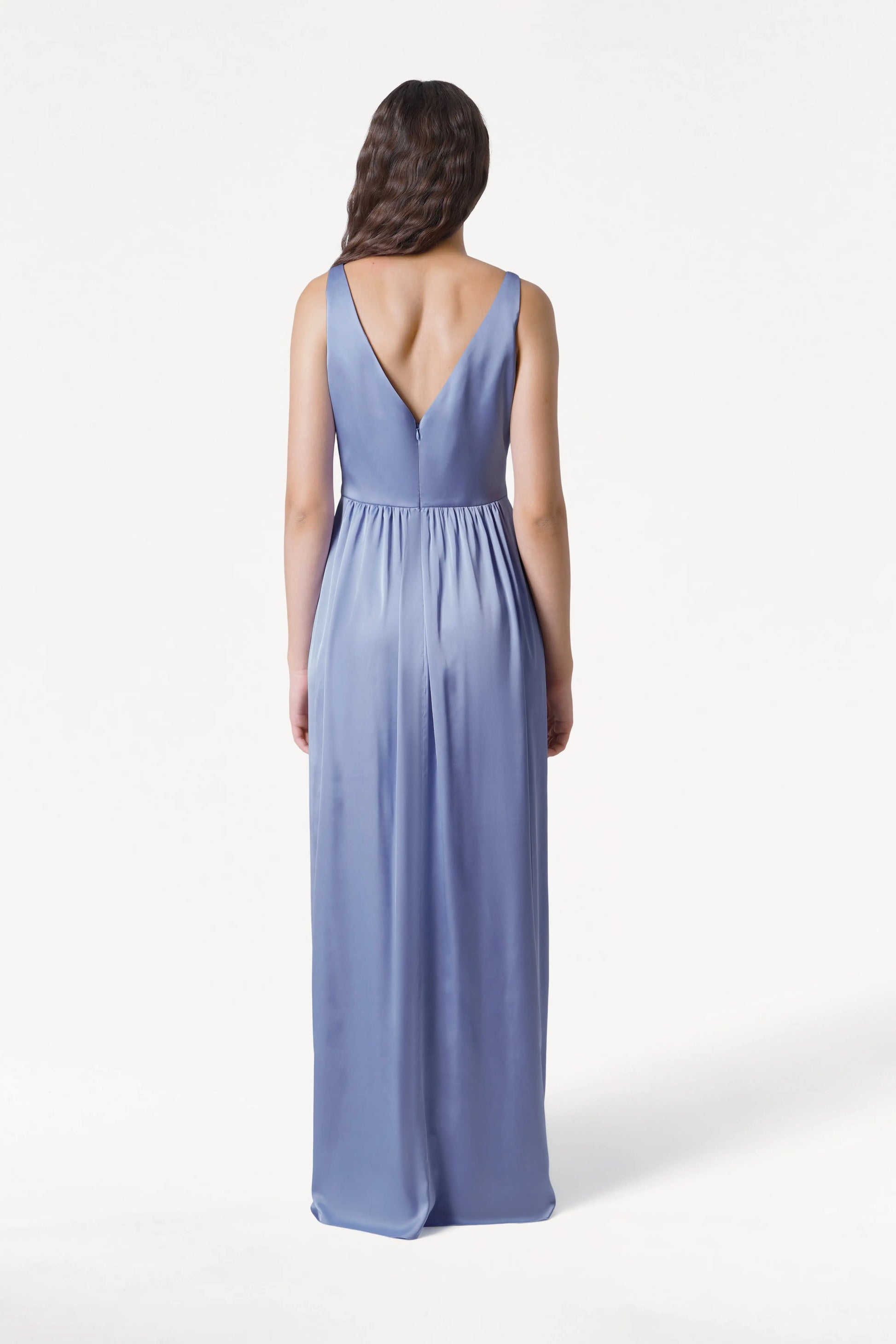 A woman with long, wavy brown hair stands facing away in the TH&TH Carmen Bridesmaid Dress in French Blue Satin, featuring a sleeveless, floor-length design and deep V-back—an elegant pick for any modern bridal party.