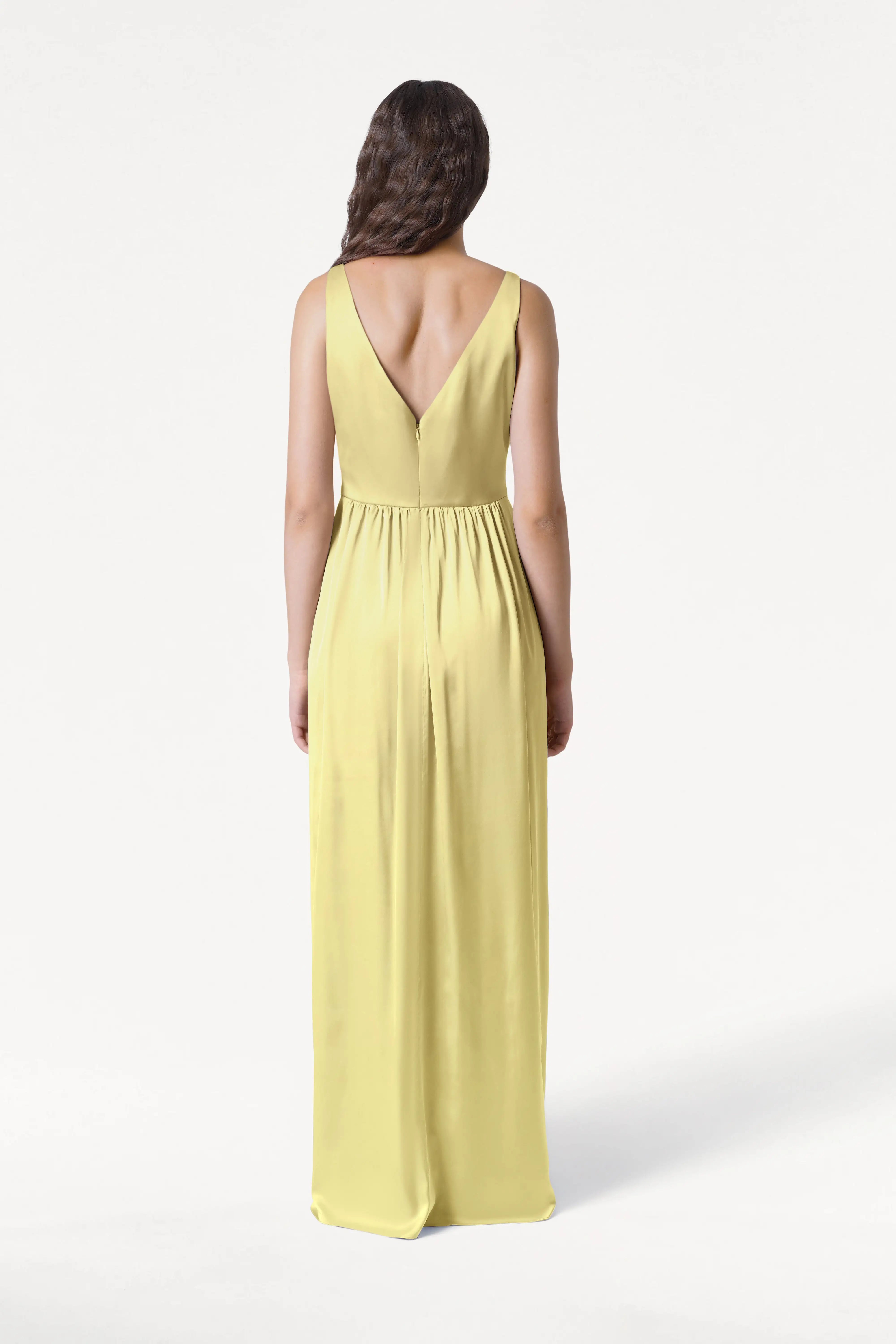 A woman with long, wavy brown hair stands with her back to the camera in the TH&TH Carmen Bridesmaid Dress in Limoncello Satin—a sleeveless, floor-length gown with a deep V-back—against a plain white background.