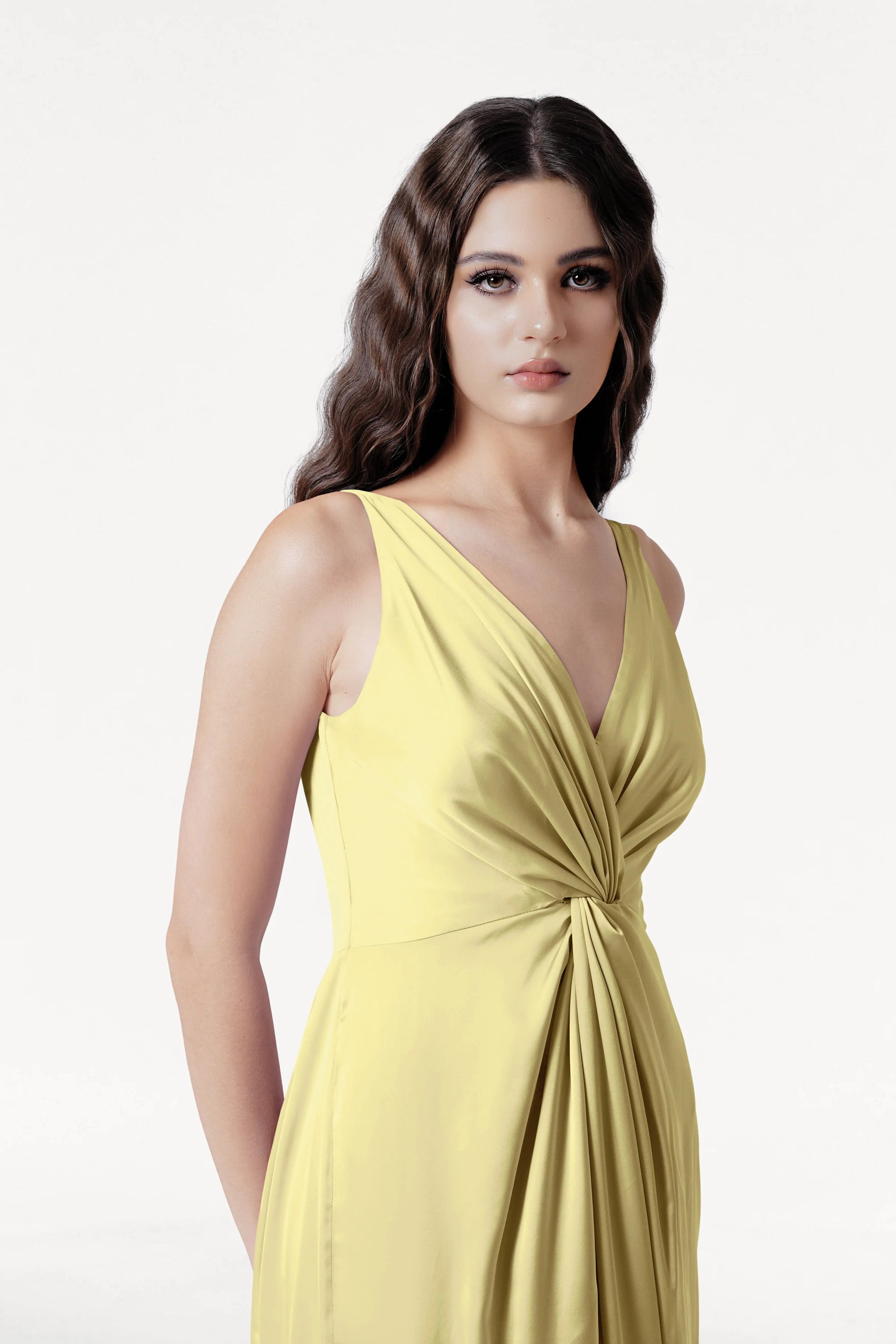 A woman with wavy brown hair models the TH&TH Carmen Bridesmaid Dress in Limoncello Satin, a sleeveless V-neck gown ideal for modern bridal parties. She stands against a plain white background, gazing neutrally at the camera.