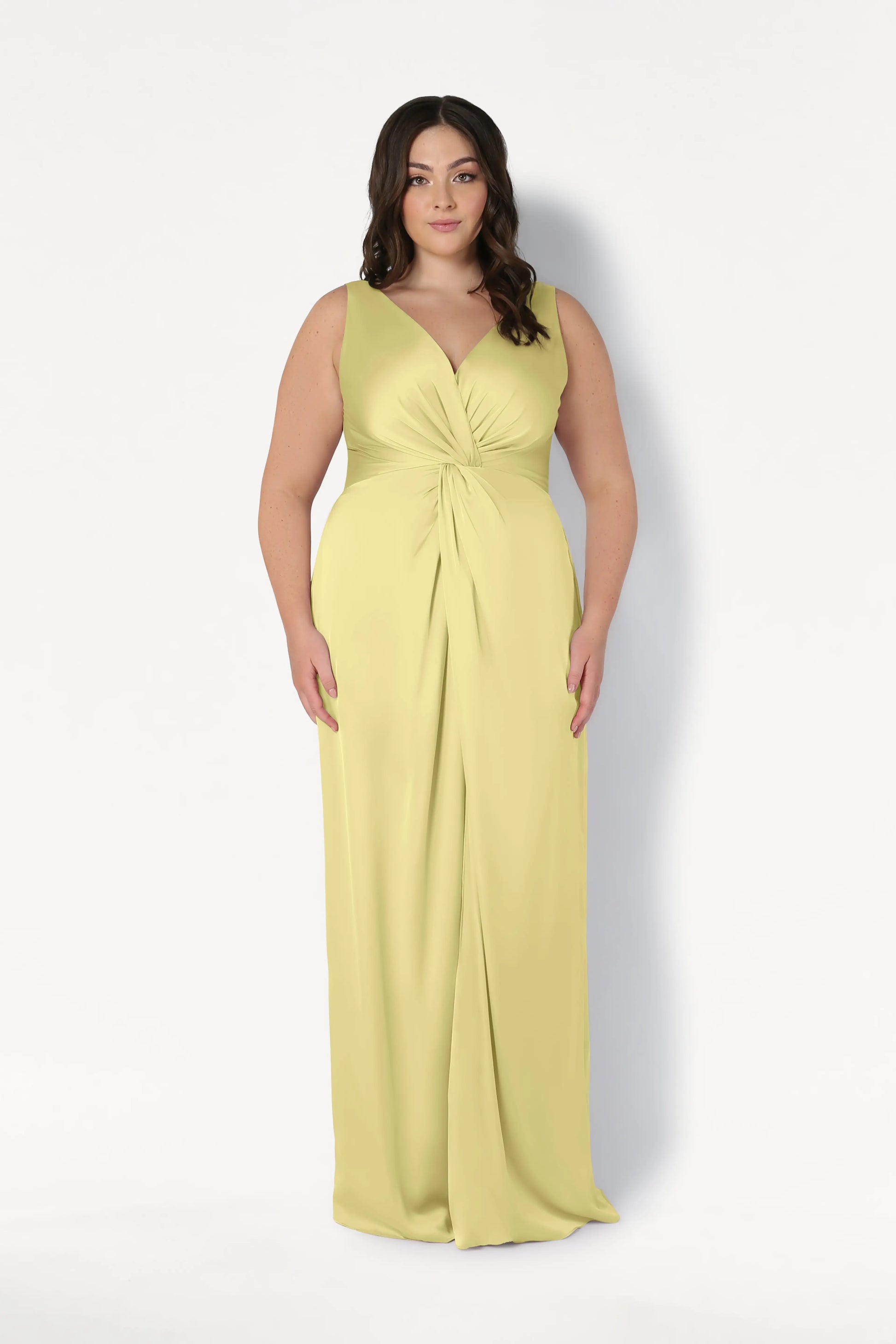 A woman with long brown hair models the TH&TH Carmen Bridesmaid Dress in Limoncello Satin—a sleeveless, floor-length gown with a V-neckline and twist-knot waist—standing against a plain white background.