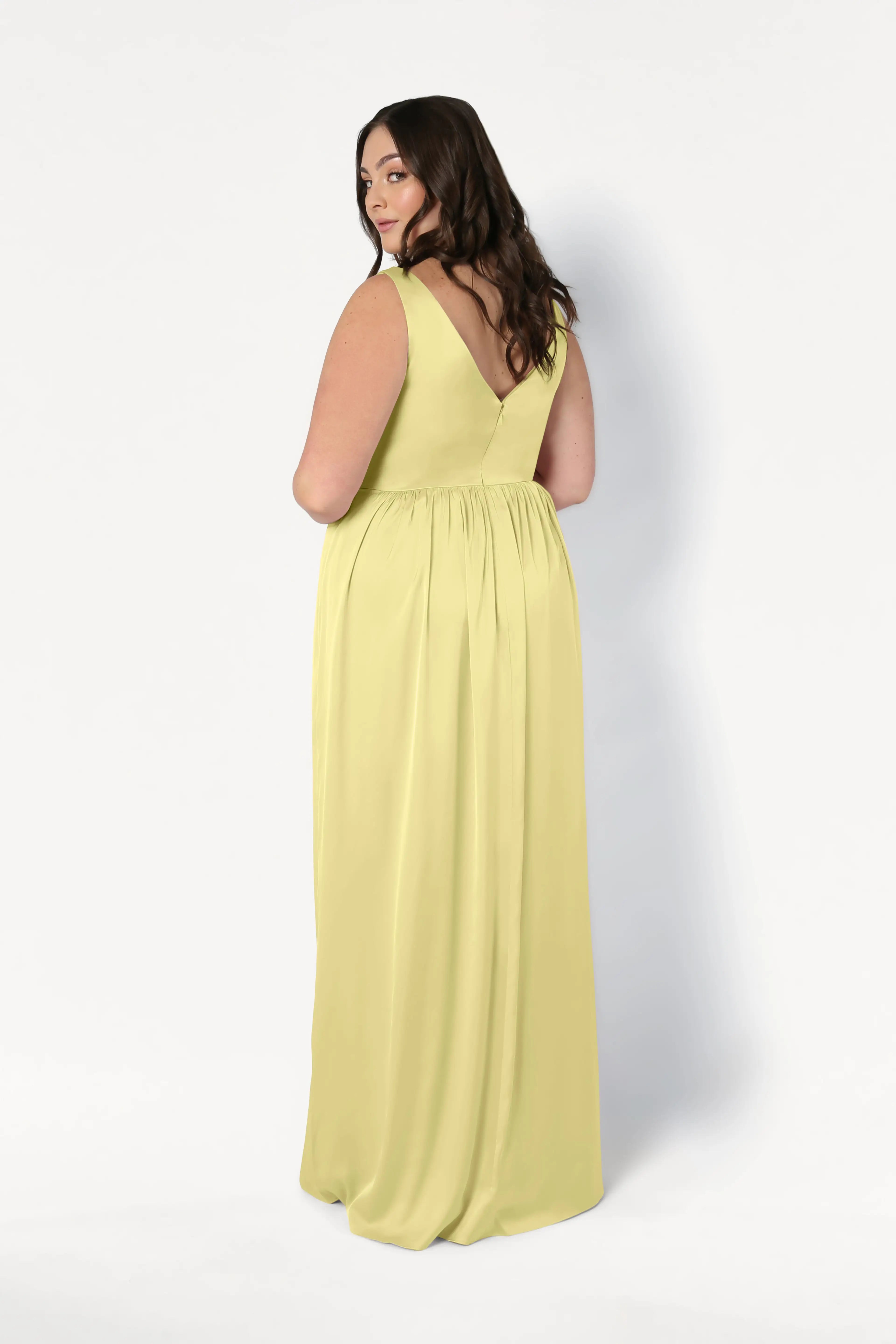 A woman with long brown hair wears the TH&TH Carmen Bridesmaid Dress in Limoncello Satin—a sleeveless, floor-length gown with a twist-knot front—posing against a plain white background. Ideal for modern bridal parties.