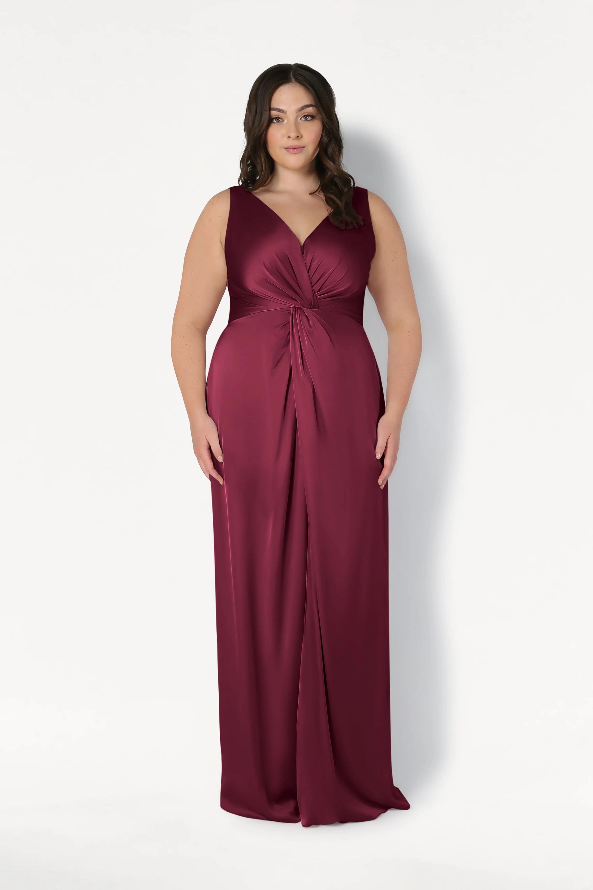 A woman with wavy dark hair wears the TH&TH Carmen Bridesmaid Dress in Merlot Satin, a sleeveless, floor-length gown with a V-neck and twist-knot waist detail, standing against a plain white background.