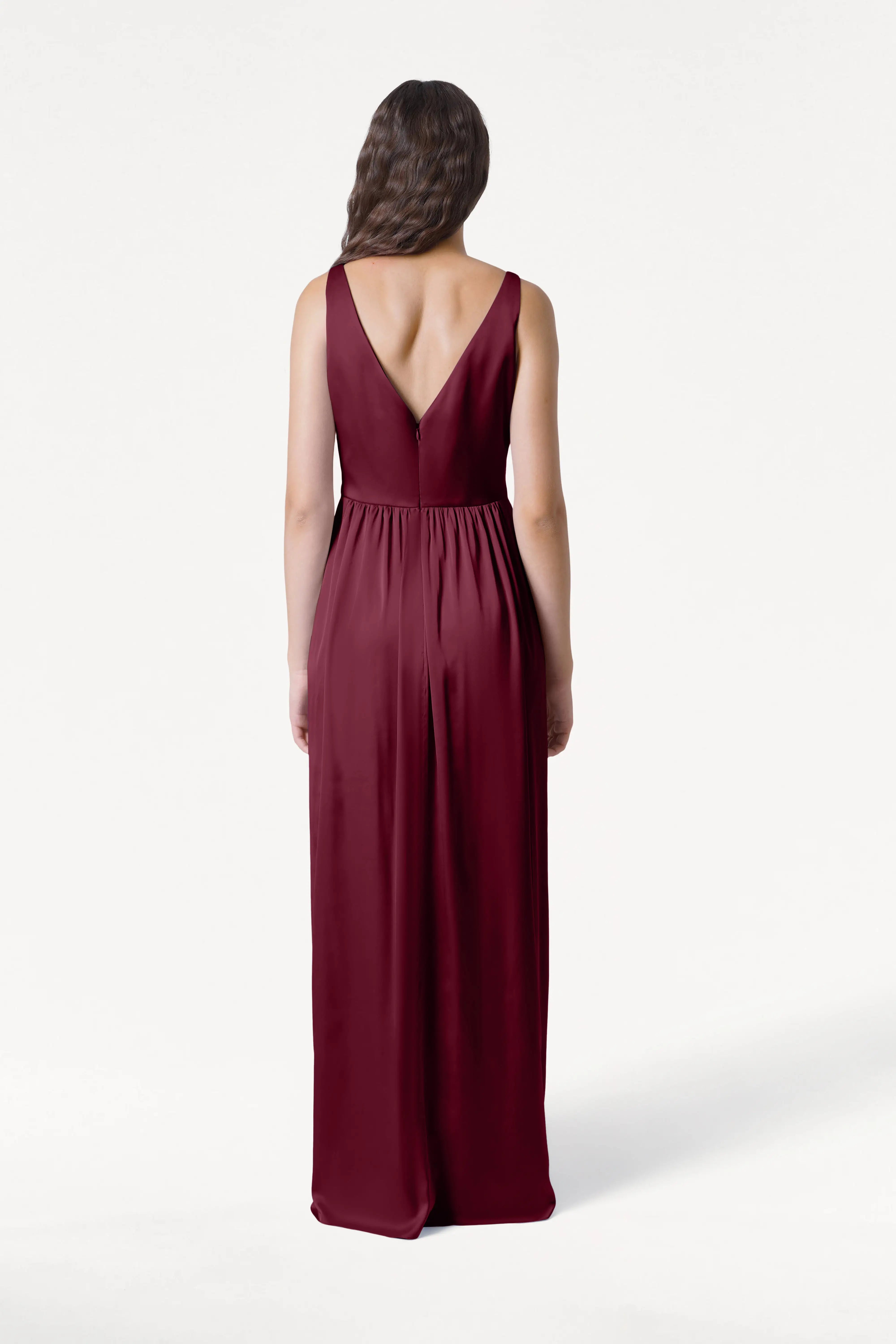 A woman with long, wavy hair stands with her back to the camera in the TH&TH Carmen Bridesmaid Dress in Merlot Satin—a sleeveless, floor-length gown with a deep V-shaped back—against a plain white background.