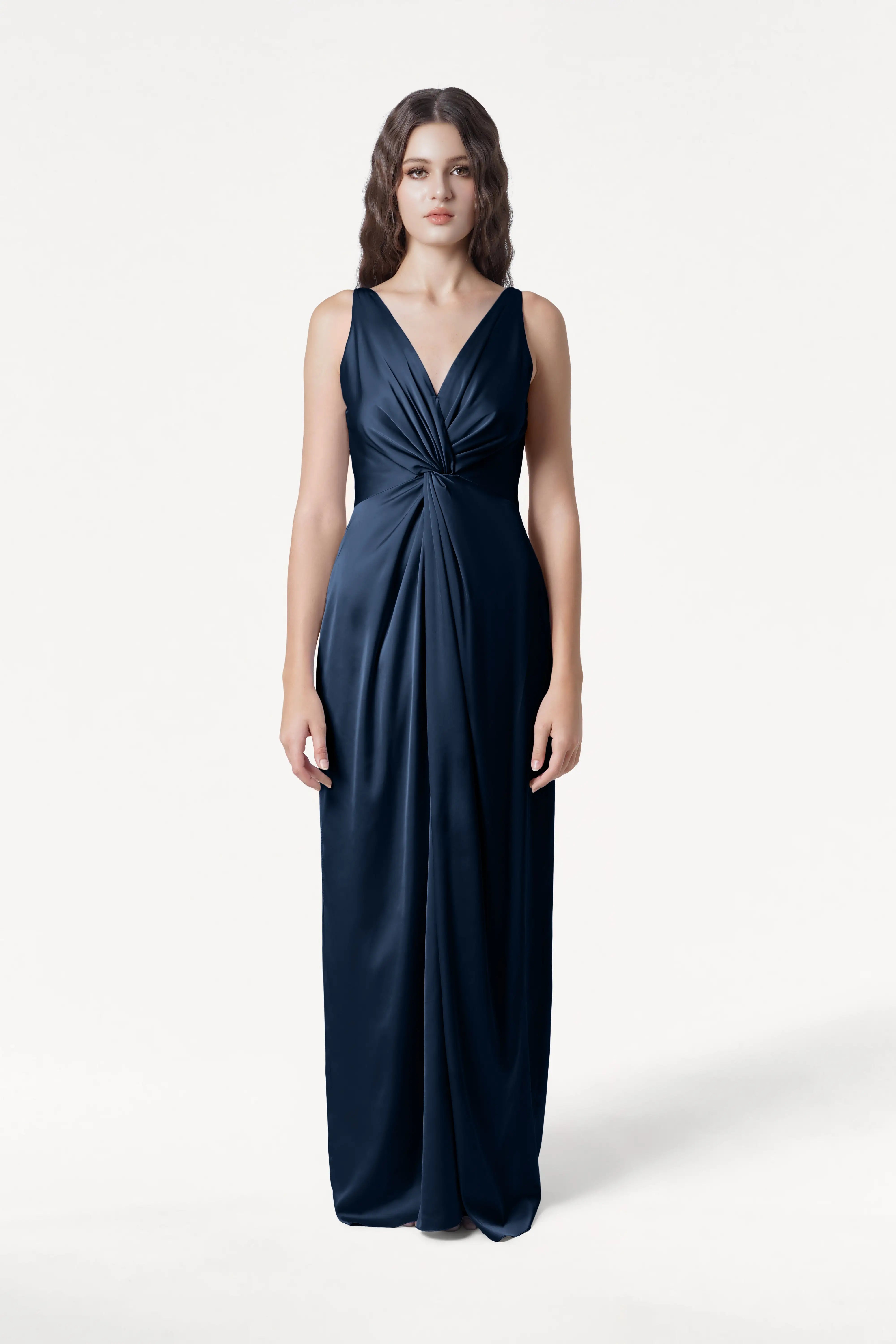 A woman with long wavy brown hair and a neutral expression faces forward, wearing the TH&TH Carmen Bridesmaid Dress in Navy Satin—a sleeveless, floor-length gown with a deep V-neck and gathered waist, perfect for modern bridal parties.