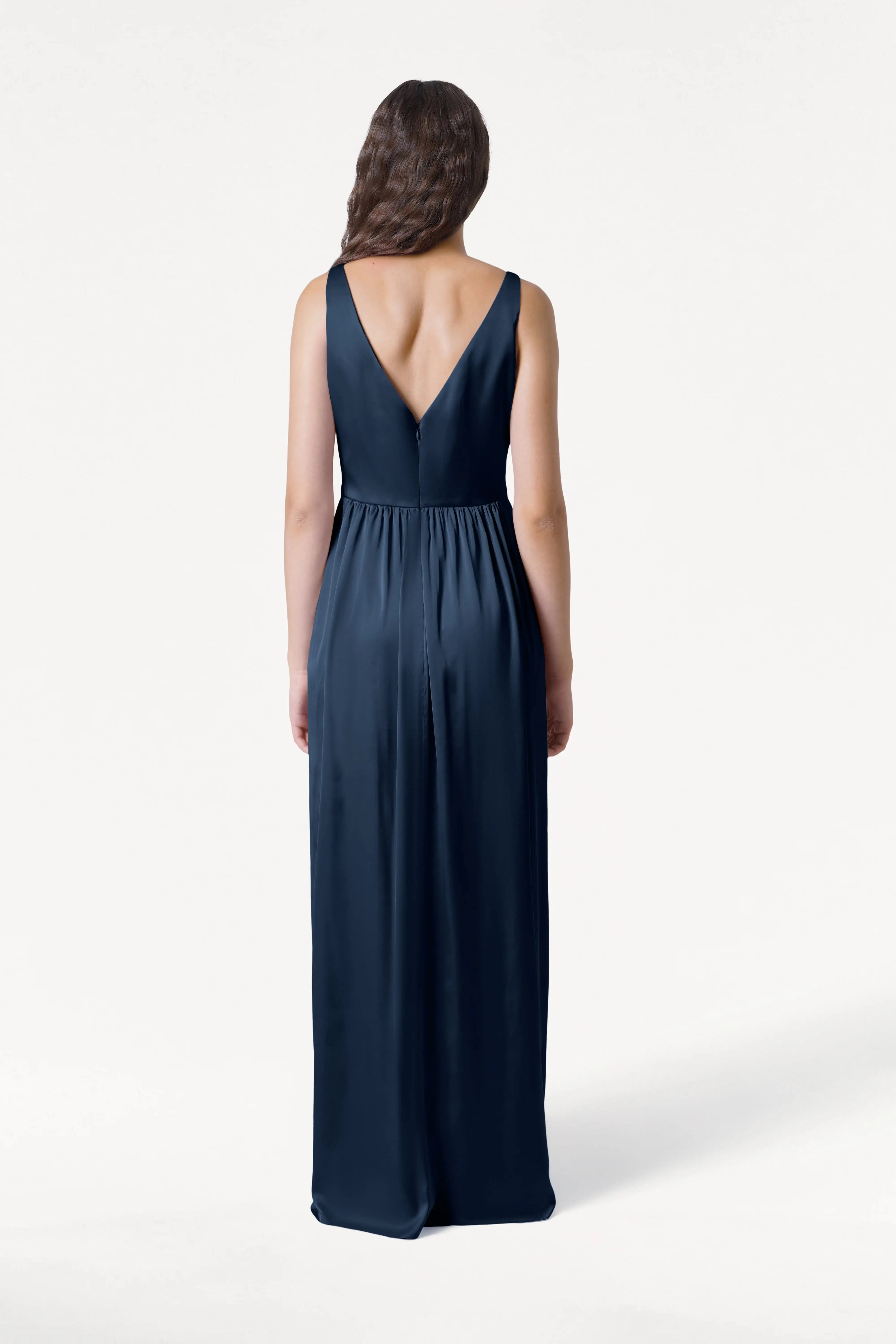 A woman with long brown hair models the TH&TH Carmen Bridesmaid Dress in Navy Satin, facing away to highlight its deep V-back and sleeveless, floor-length design—an elegant choice for modern bridal parties, shown against a plain white background.