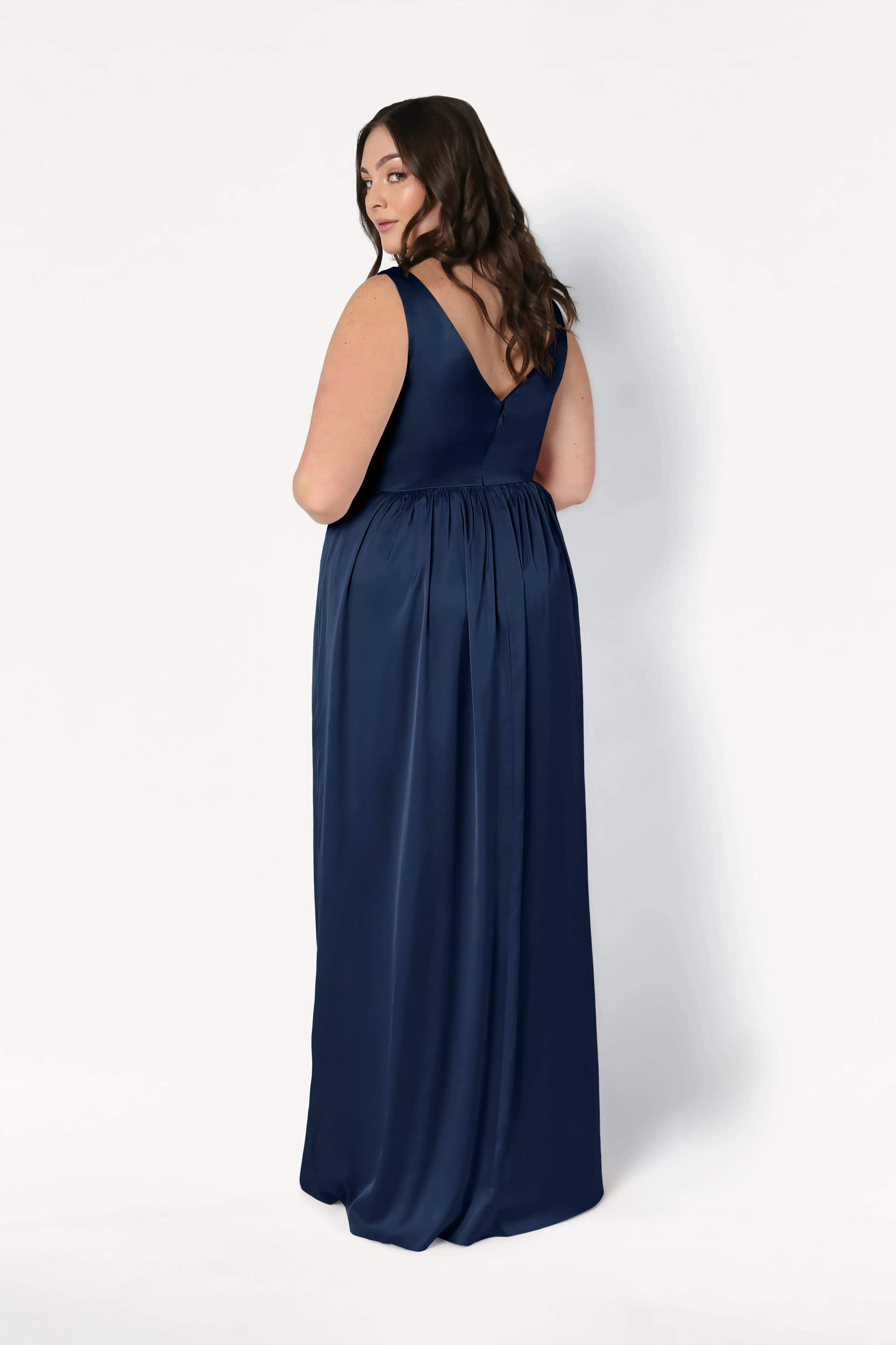 A woman with long brown hair poses backward, looking over her shoulder in the TH&TH Carmen Bridesmaid Dress in Navy Satin—a sleeveless, floor-length gown with a deep V-back—perfect for modern bridal parties against a plain white background.
