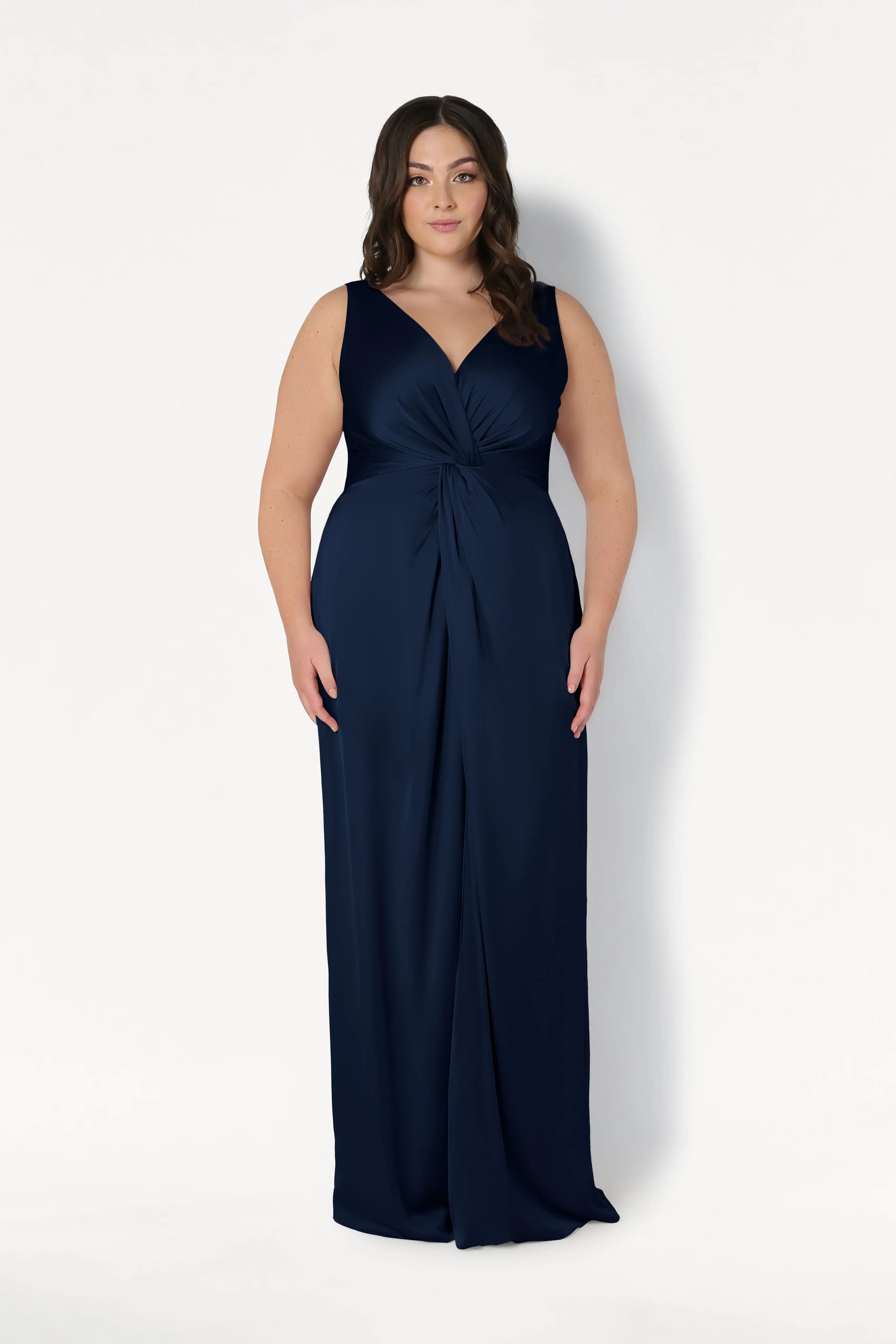 A woman with long brown hair models the TH&TH Carmen Bridesmaid Dress in Navy Satin, featuring a V-neckline and gathered knot waist detail, perfect for modern bridal parties, standing against a plain white background.