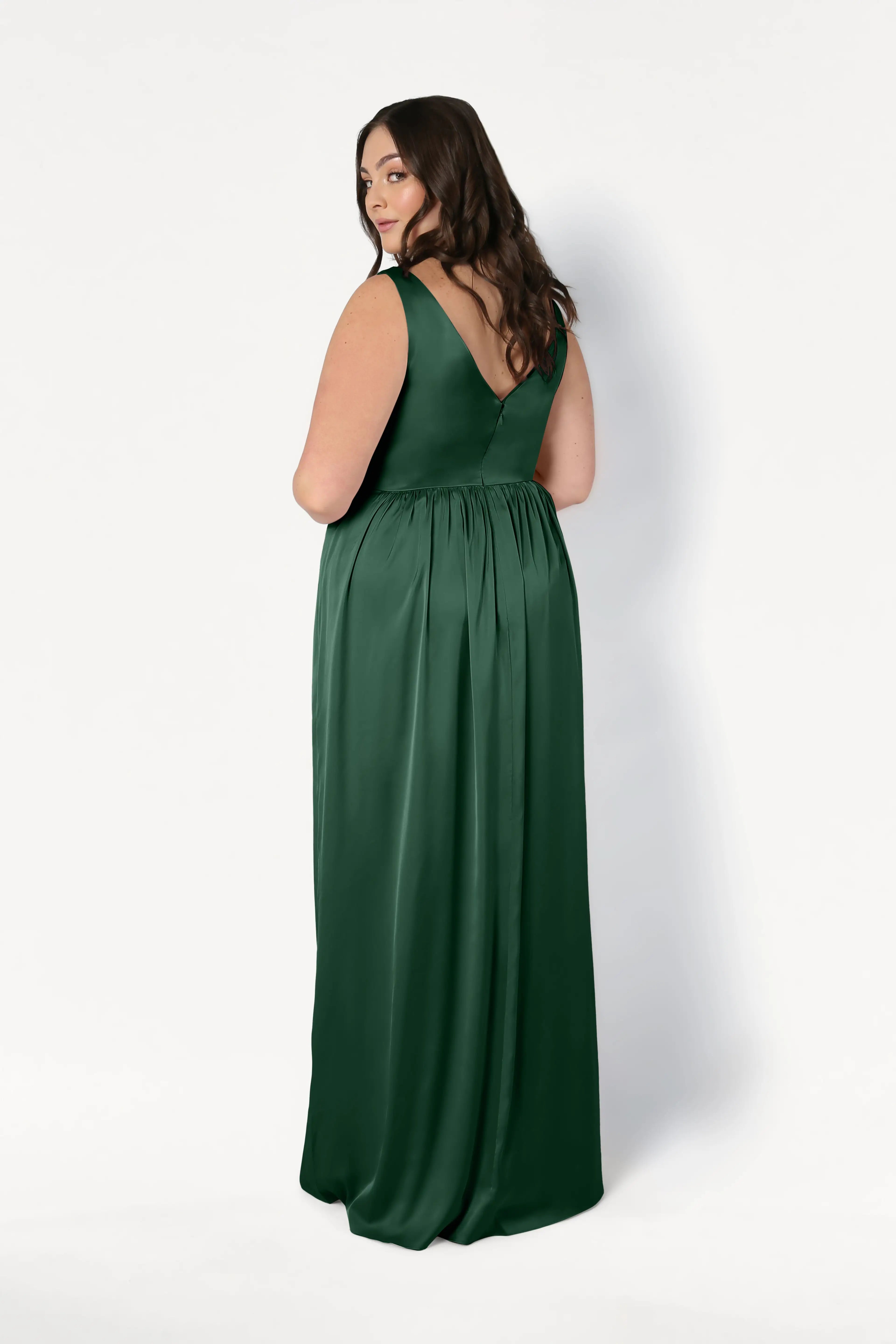 A woman in a sleeveless, floor-length Carmen Bridesmaid Dress in Olive Grove Satin by TH&TH stands with a deep V-back, glancing over her shoulder against a white background—ideal for modern bridal parties.