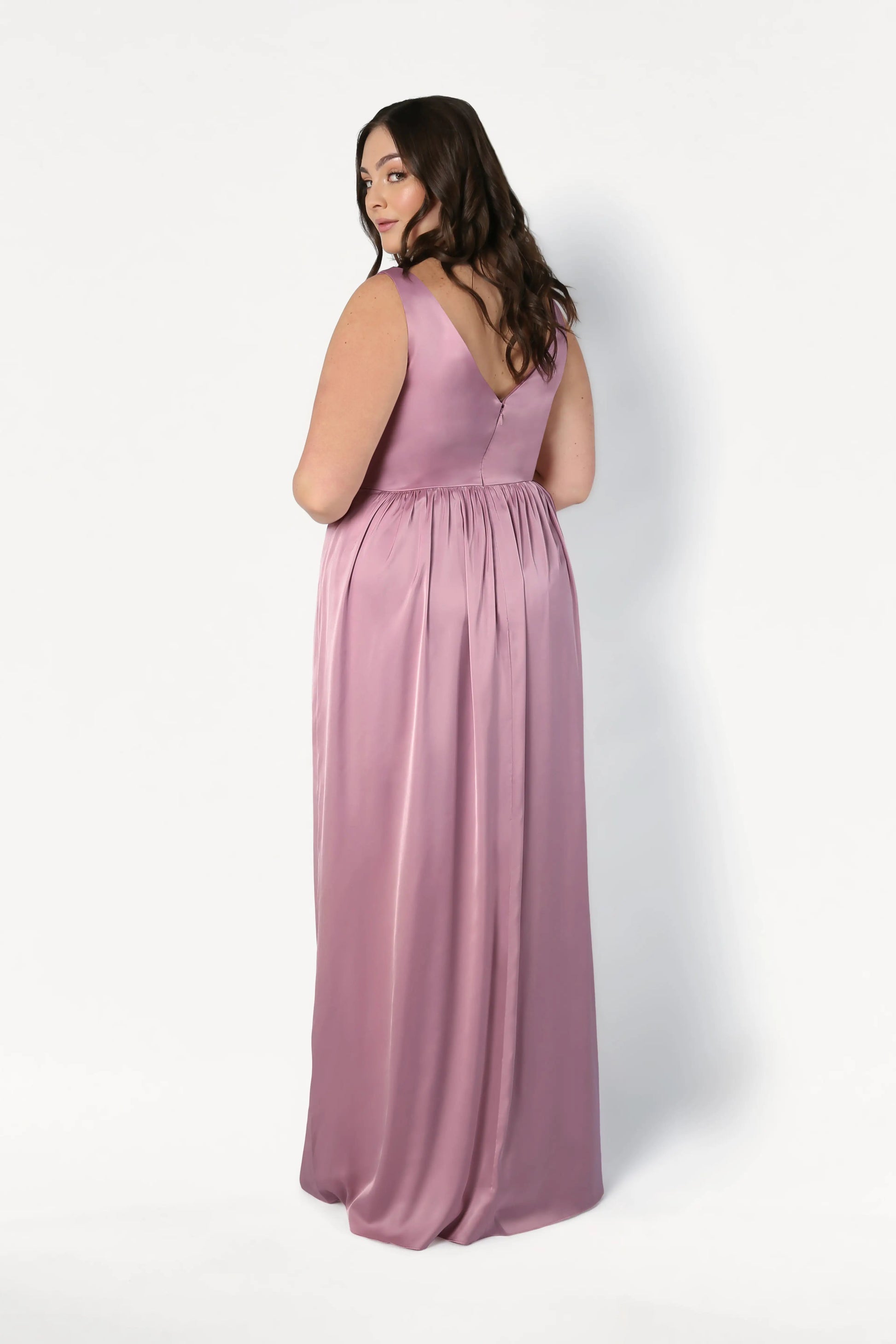 A woman with long brown hair looks over her shoulder, wearing the TH&TH Carmen Bridesmaid Dress in rose pink satin—a sleeveless, floor-length gown with a V-shaped back—set against a plain white background.