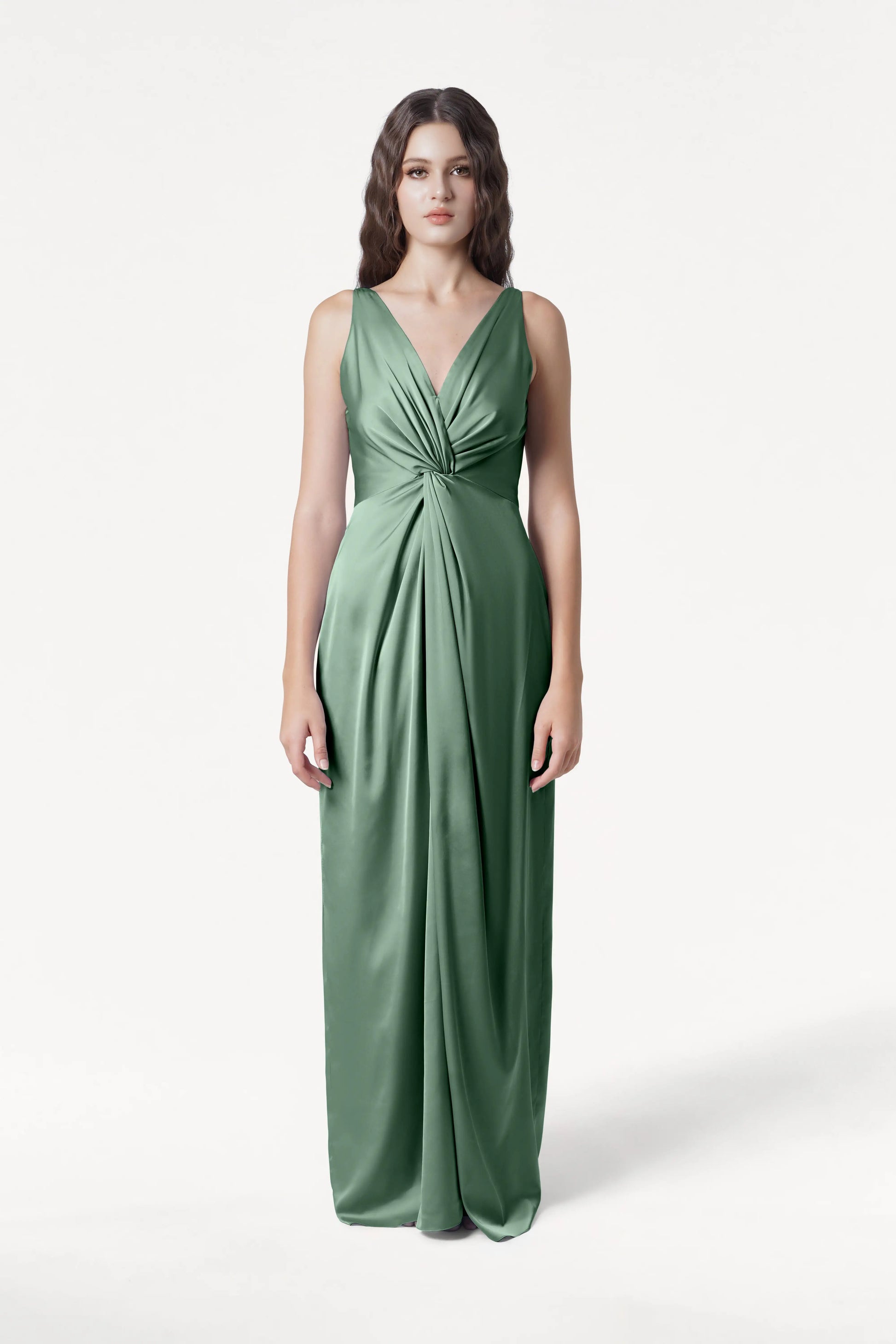 A woman faces forward in the TH&TH Carmen Bridesmaid Dress, featuring sage green satin and a twist-knot detail at the waist. She has long, wavy brown hair and a neutral expression, standing against a plain white background—a chic choice for bridal parties.