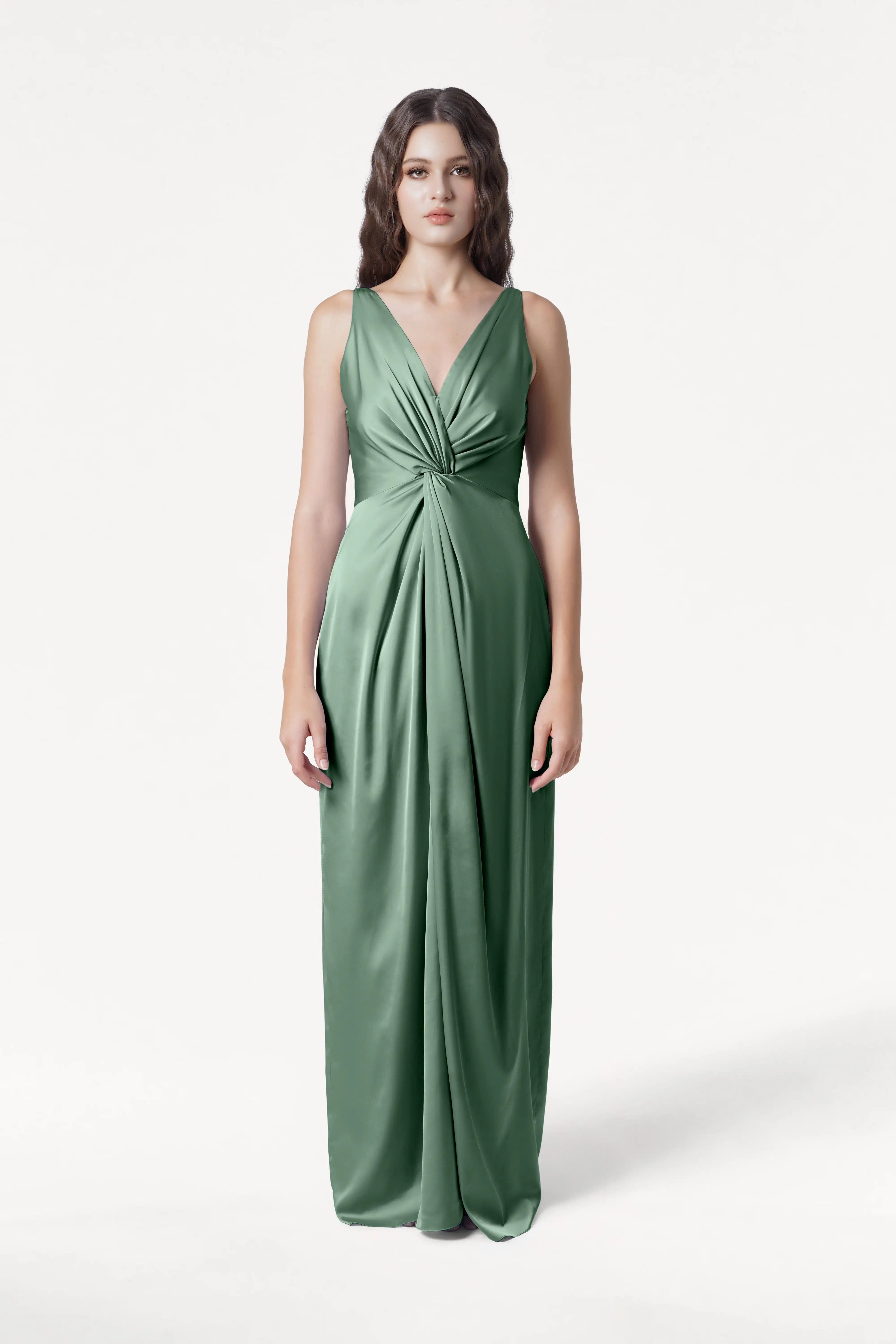 A woman faces forward in the TH&TH Carmen Bridesmaid Dress, featuring sage green satin and a twist-knot detail at the waist. She has long, wavy brown hair and a neutral expression, standing against a plain white background—a chic choice for bridal parties.
