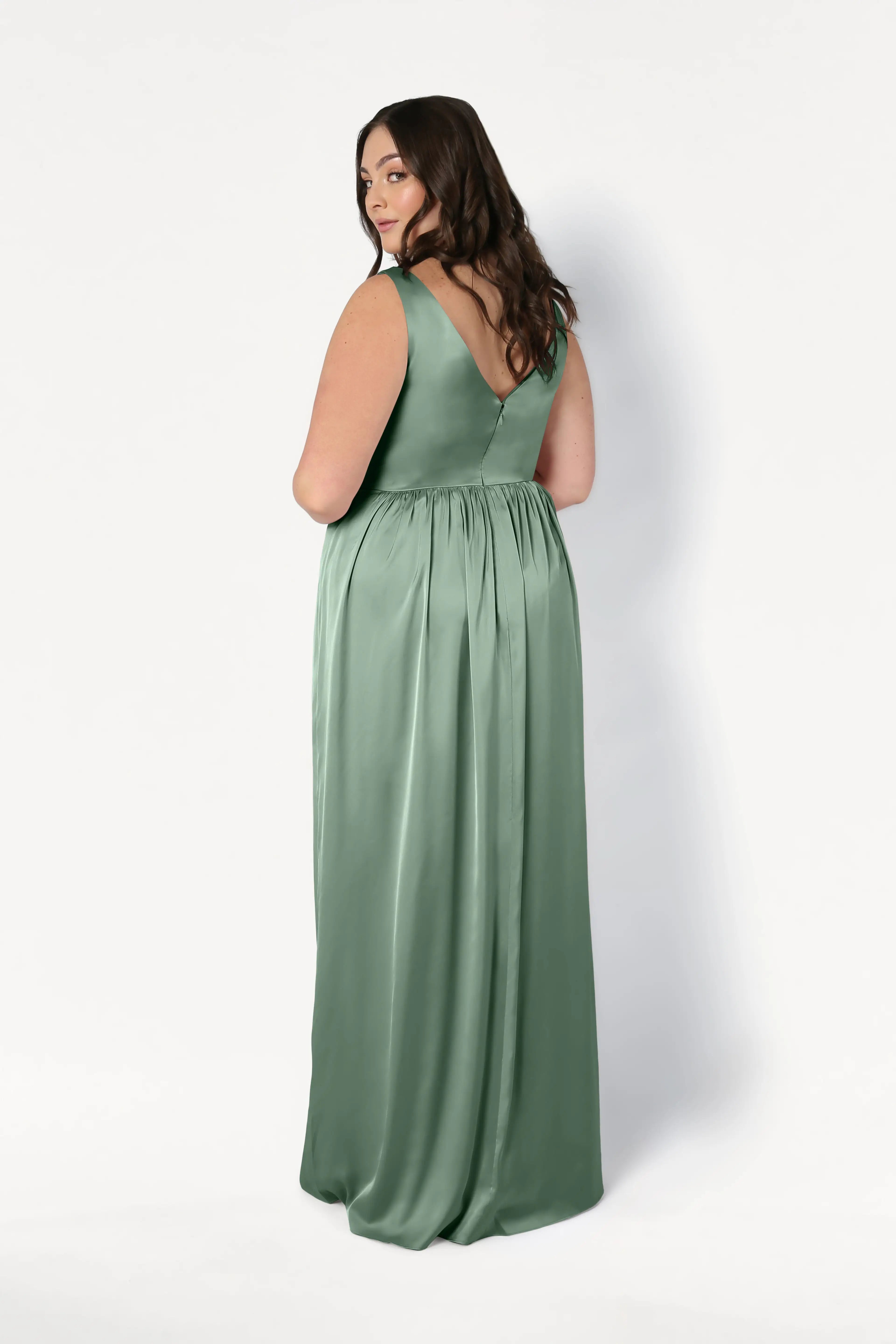 A woman with long brown hair wears the TH&TH Carmen Bridesmaid Dress in Sage Green Satin. Sleeveless and floor-length, she faces away but looks over her shoulder—a modern, flattering choice for bridal parties.