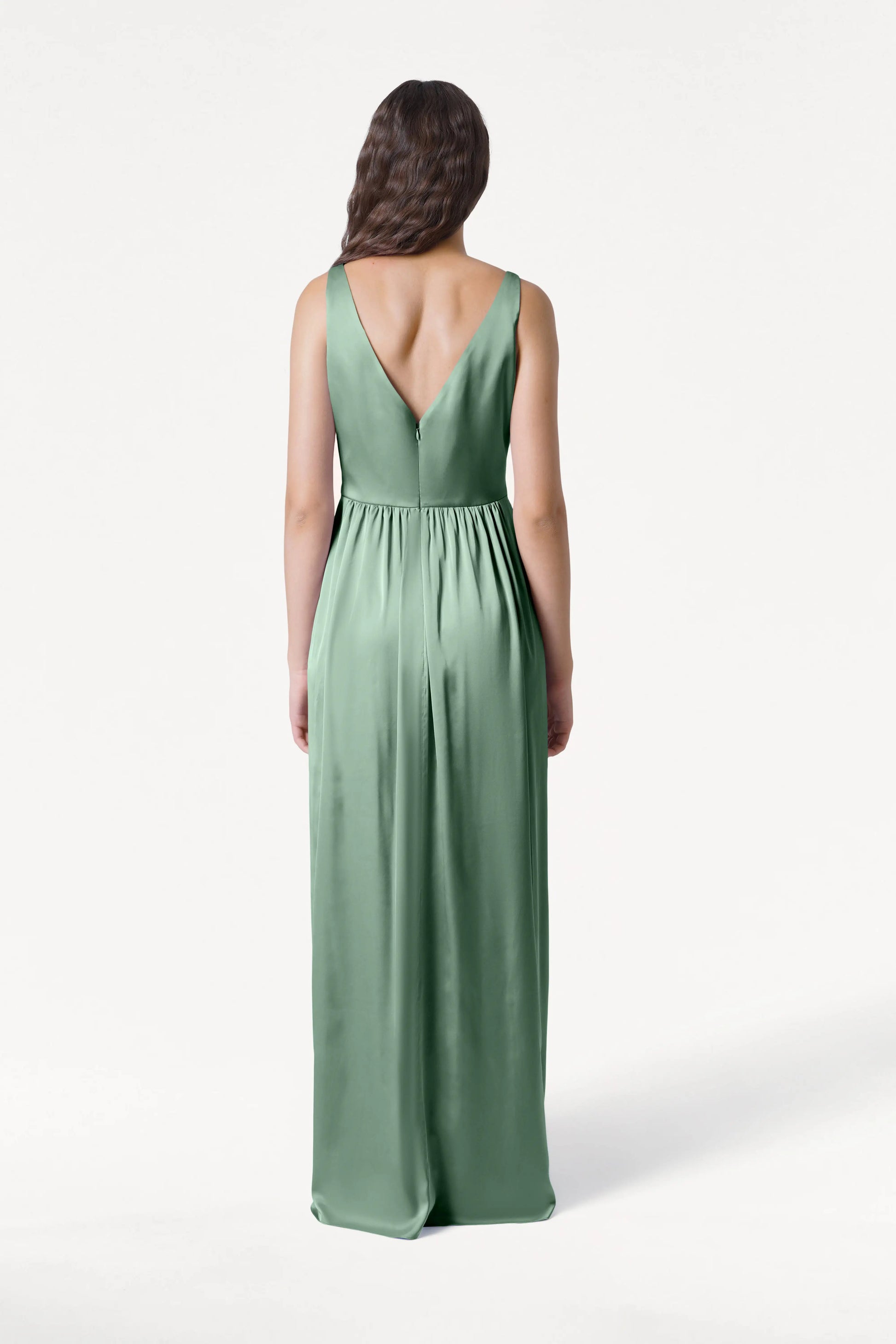 A woman with long wavy brown hair faces away, modeling the Carmen Bridesmaid Dress in Sage Green Satin by TH&TH—a sleeveless, floor-length gown with a deep V-back and gathered waist—set against a plain white background.