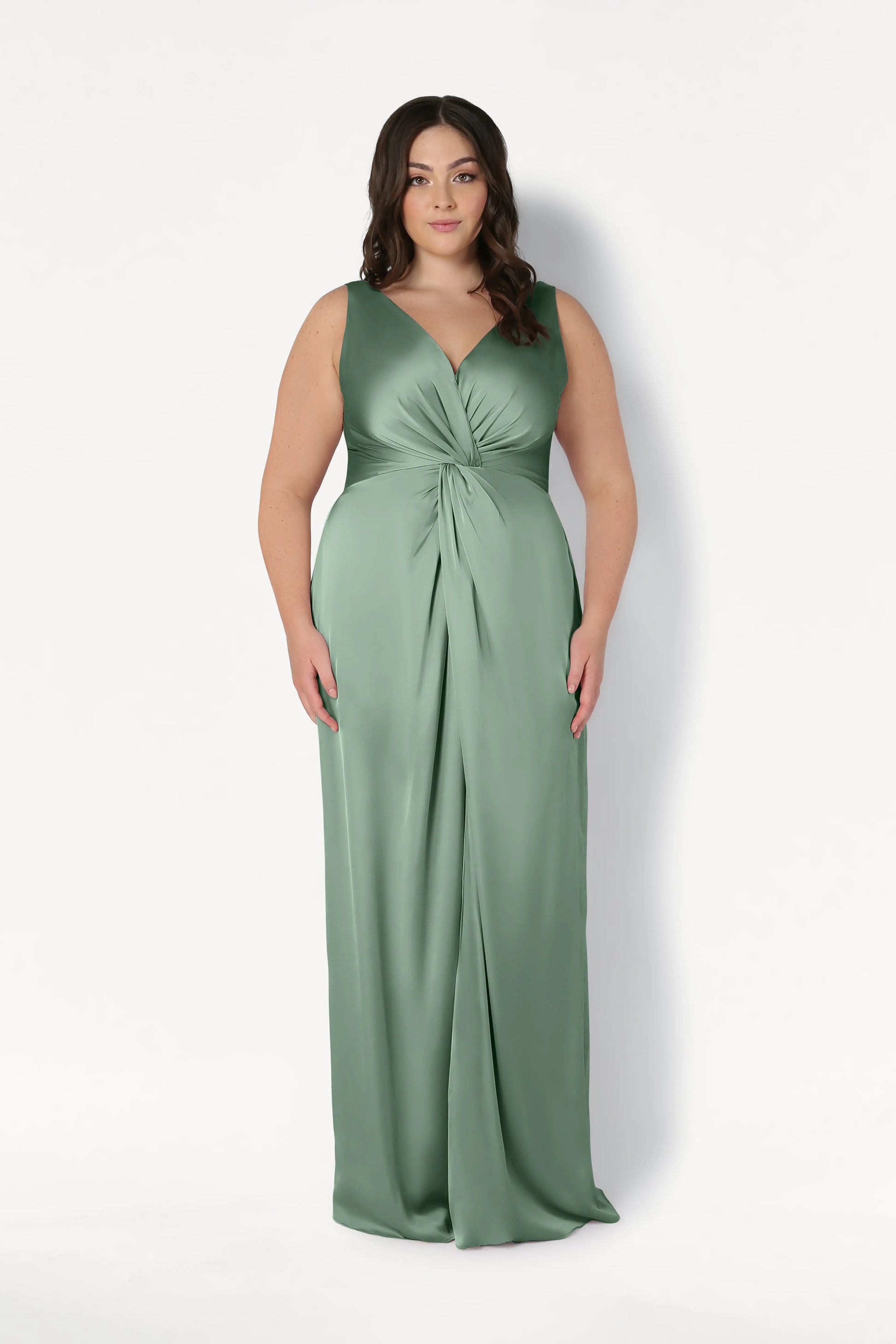 A woman models the TH&TH Carmen Bridesmaid Dress in Sage Green Satin—sleeveless, floor-length with a twist-knot front. She stands against a white background, her long wavy brown hair and neutral expression suiting modern bridal parties.