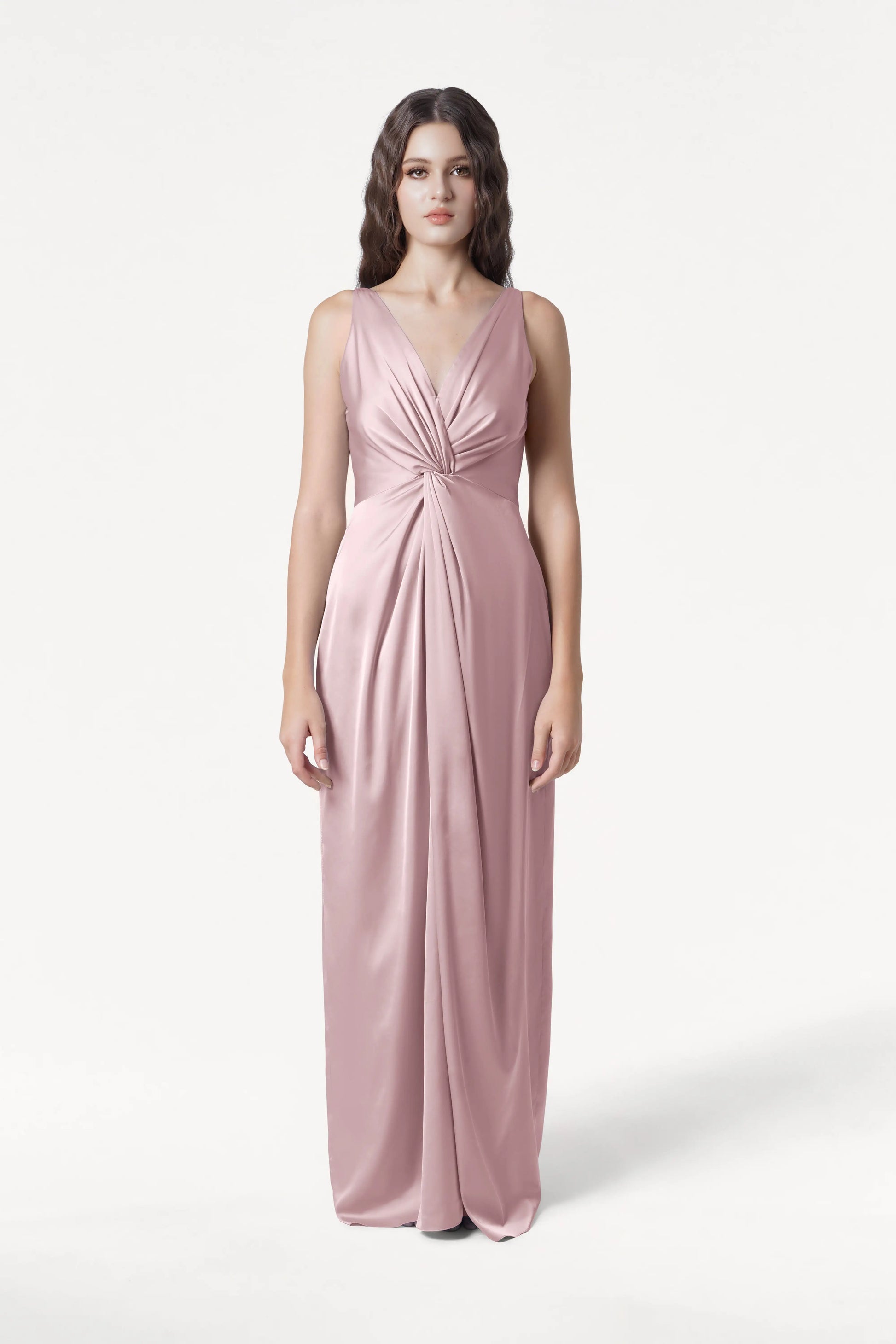 A woman poses against a white background in the TH&TH Carmen Bridesmaid Dress—an elegant, sleeveless, floor-length smoked blush satin gown with a deep V-neckline and gathered waist, ideal for modern bridal parties.
