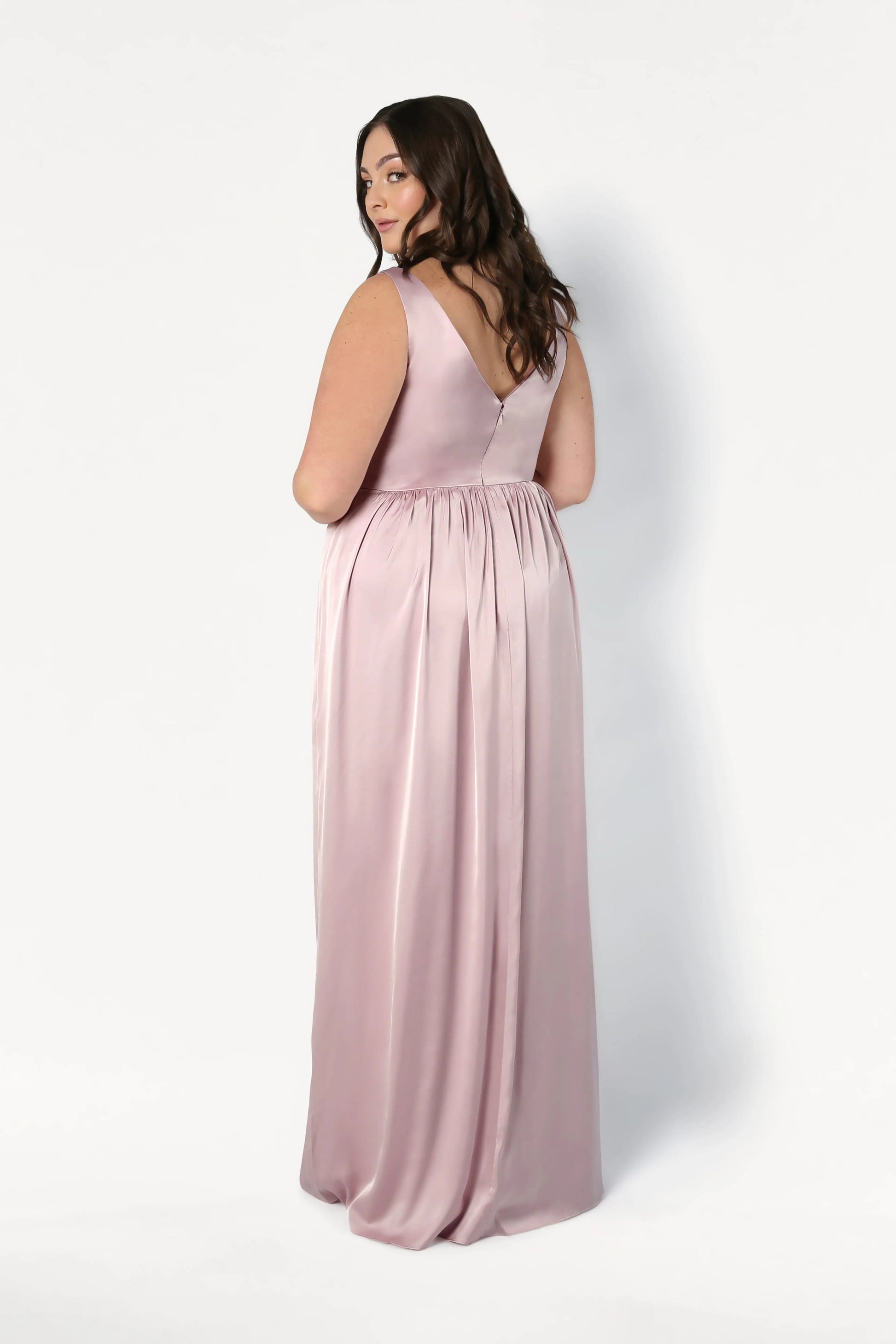 A woman with long brown hair models the TH&TH Carmen Bridesmaid Dress in Smoked Blush Satin—a sleeveless, floor-length gown—standing against a white background and looking over her shoulder; a chic choice for modern bridal parties.