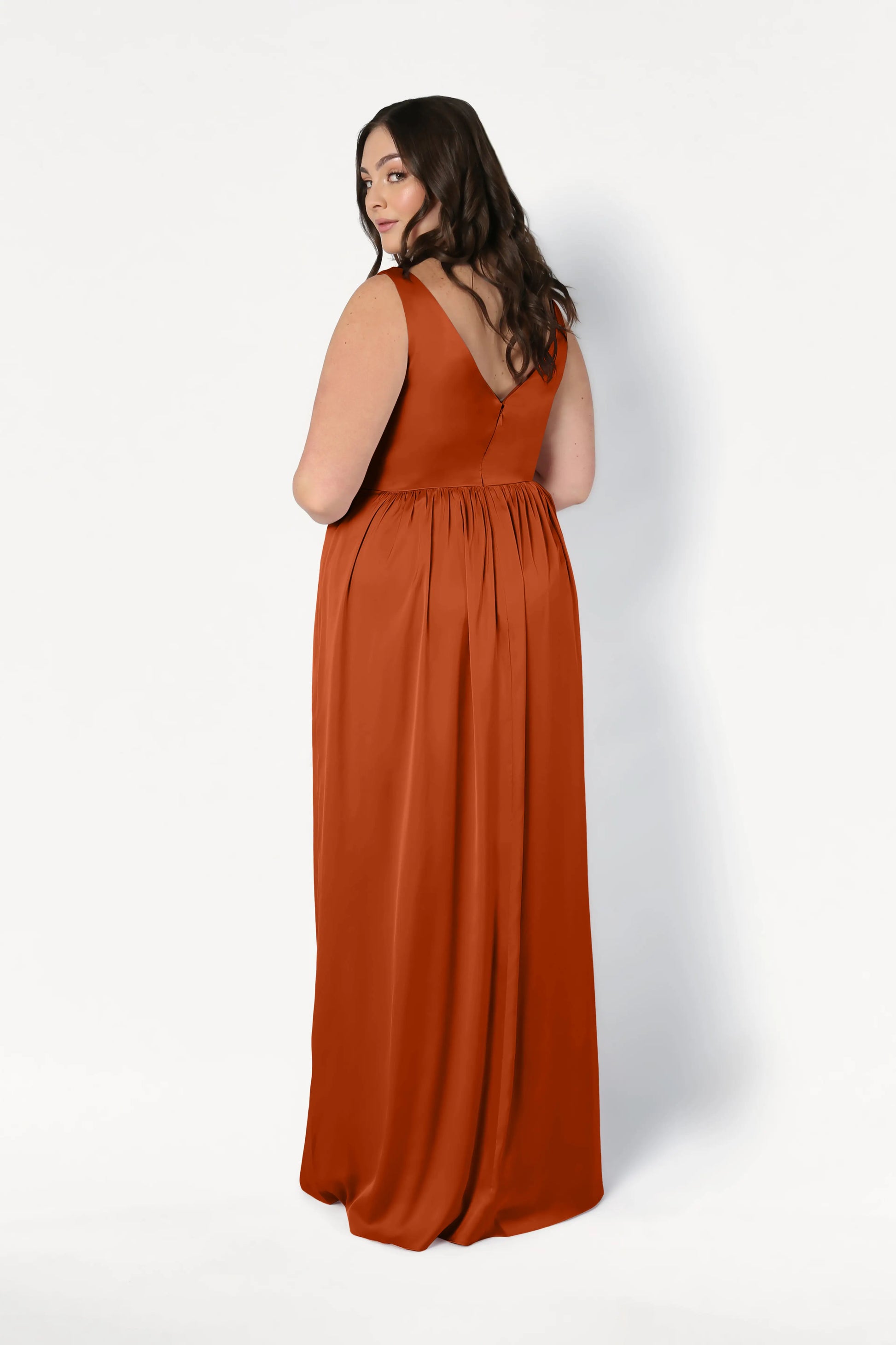 A woman with long brown hair wears the TH&TH Carmen Bridesmaid Dress in Terracotta Satin, a sleeveless, floor-length gown, standing against a plain white background—ideal for modern bridal parties.