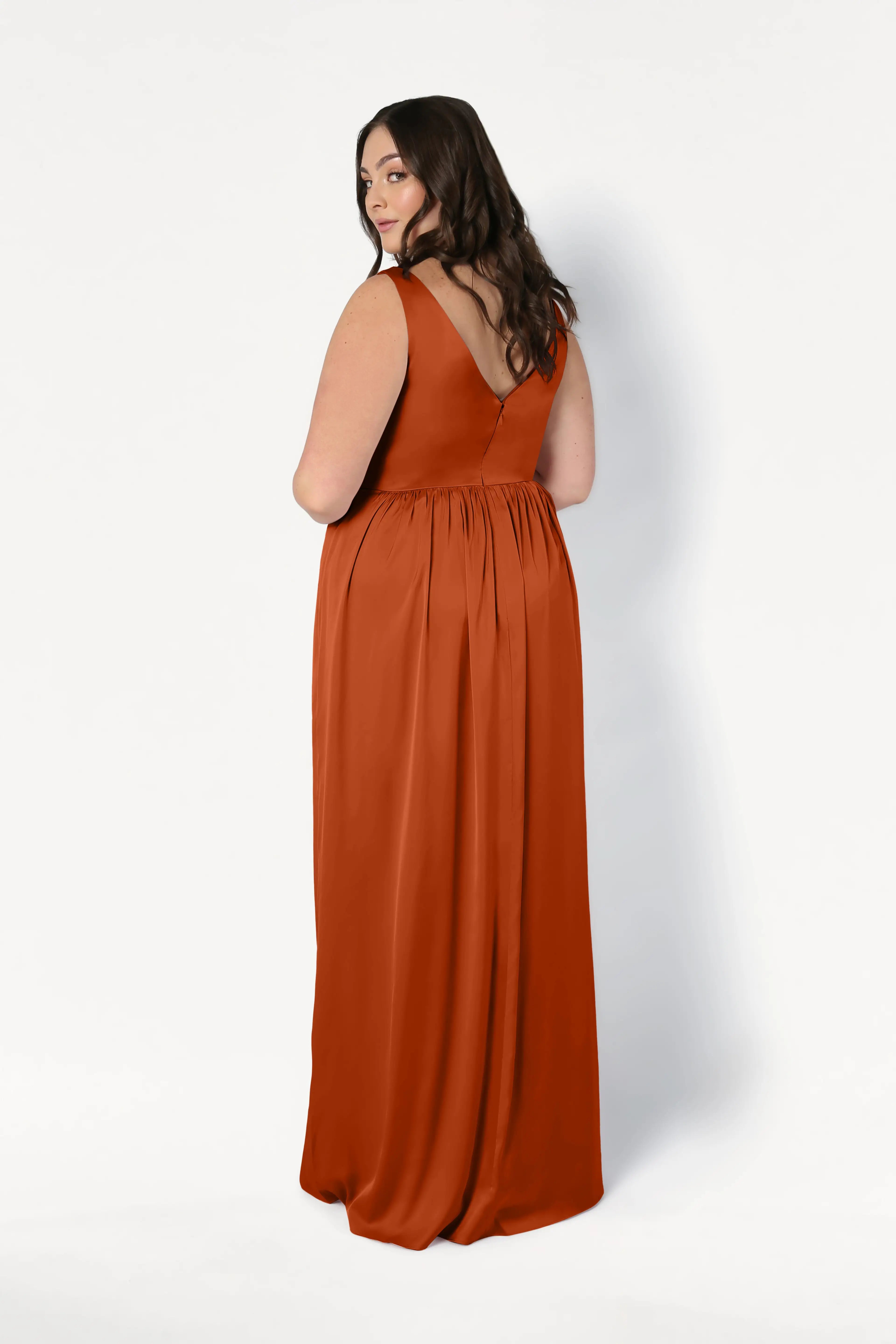 A woman with long brown hair wears the TH&TH Carmen Bridesmaid Dress in Terracotta Satin, a sleeveless, floor-length gown, standing against a plain white background—ideal for modern bridal parties.