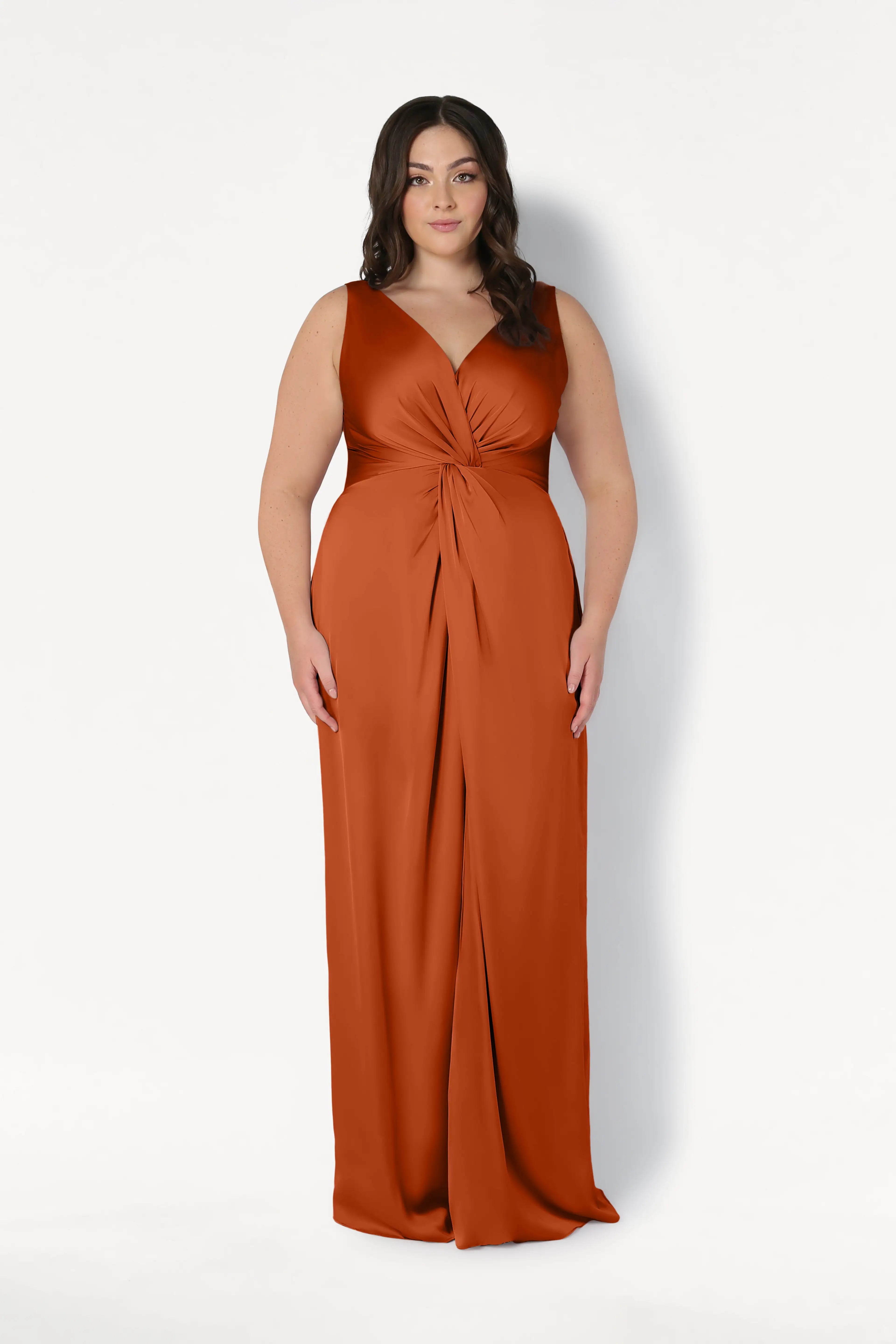 A woman wears the Carmen Bridesmaid Dress in Terracotta Satin by TH&TH—an elegant, sleeveless V-neck gown with a gathered twist waist detail—perfect for modern bridal parties, shown against a plain white background.