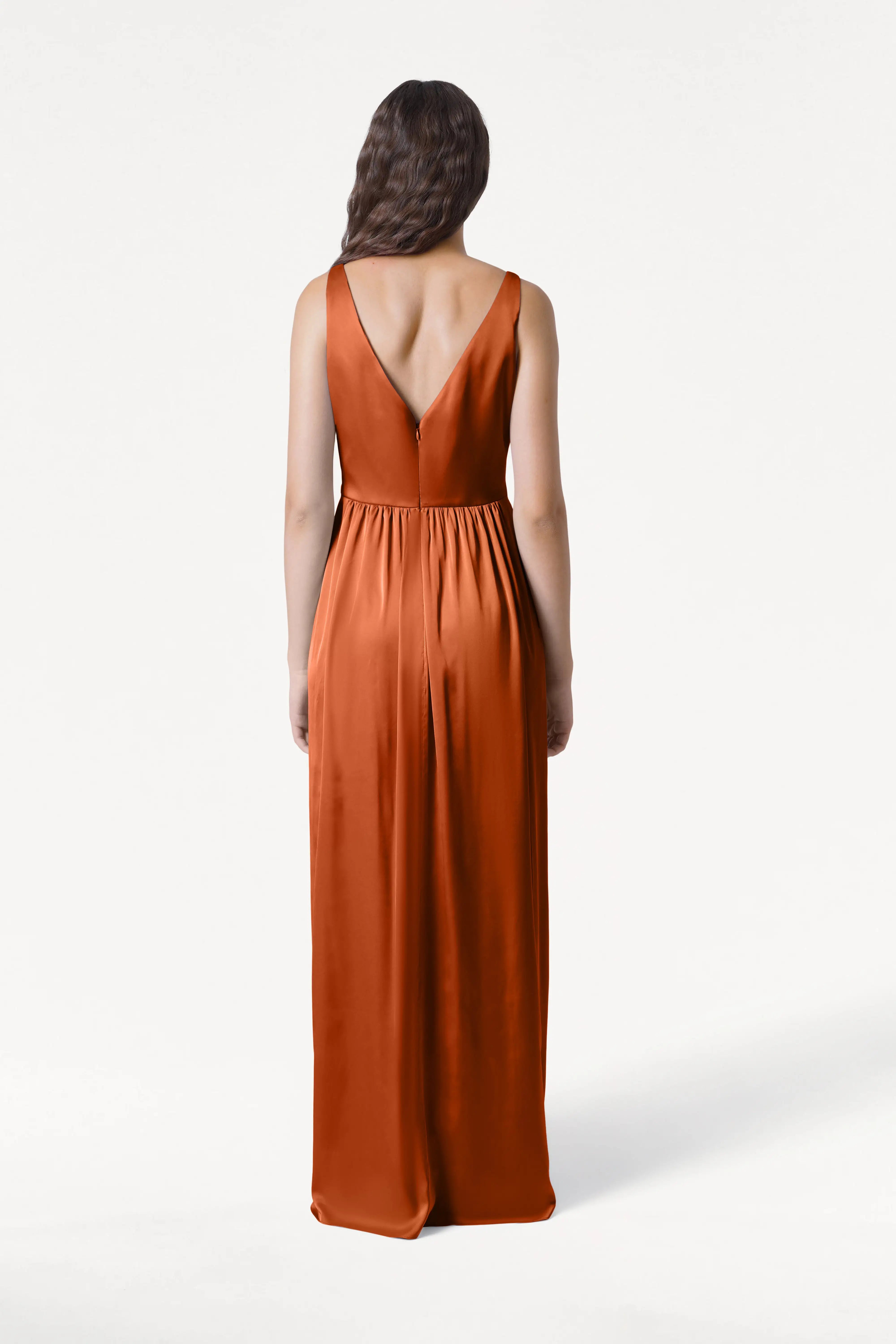 A woman stands with her back to the camera, modeling the TH&TH Carmen Bridesmaid Dress in Terracotta Satin—a floor-length, sleeveless gown with a deep V-back and gathered waist—set against a plain white background.