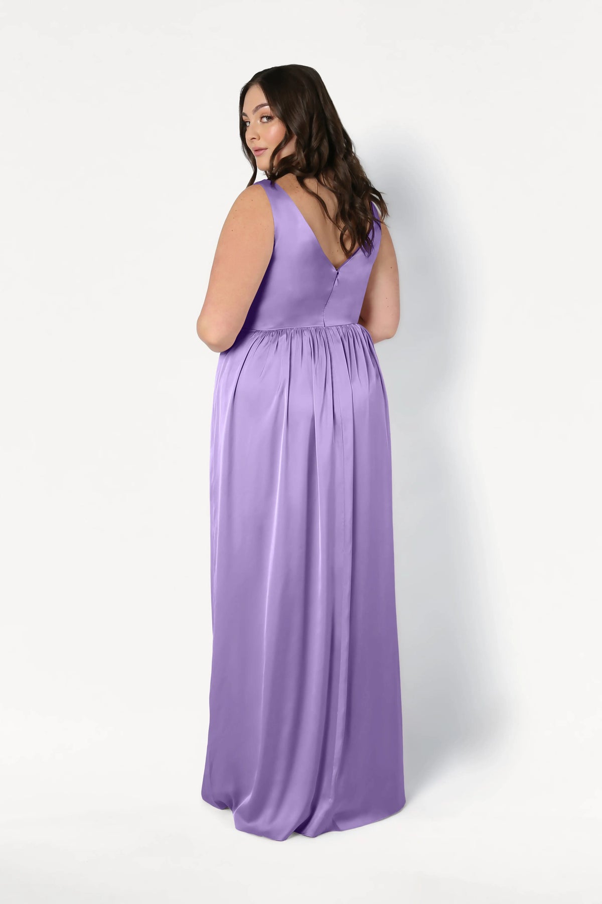 A woman with long brown hair poses with her back to the camera, showcasing the TH&TH Carmen Bridesmaid Dress in Violet Satin, a sleeveless floor-length gown, against a plain white background.