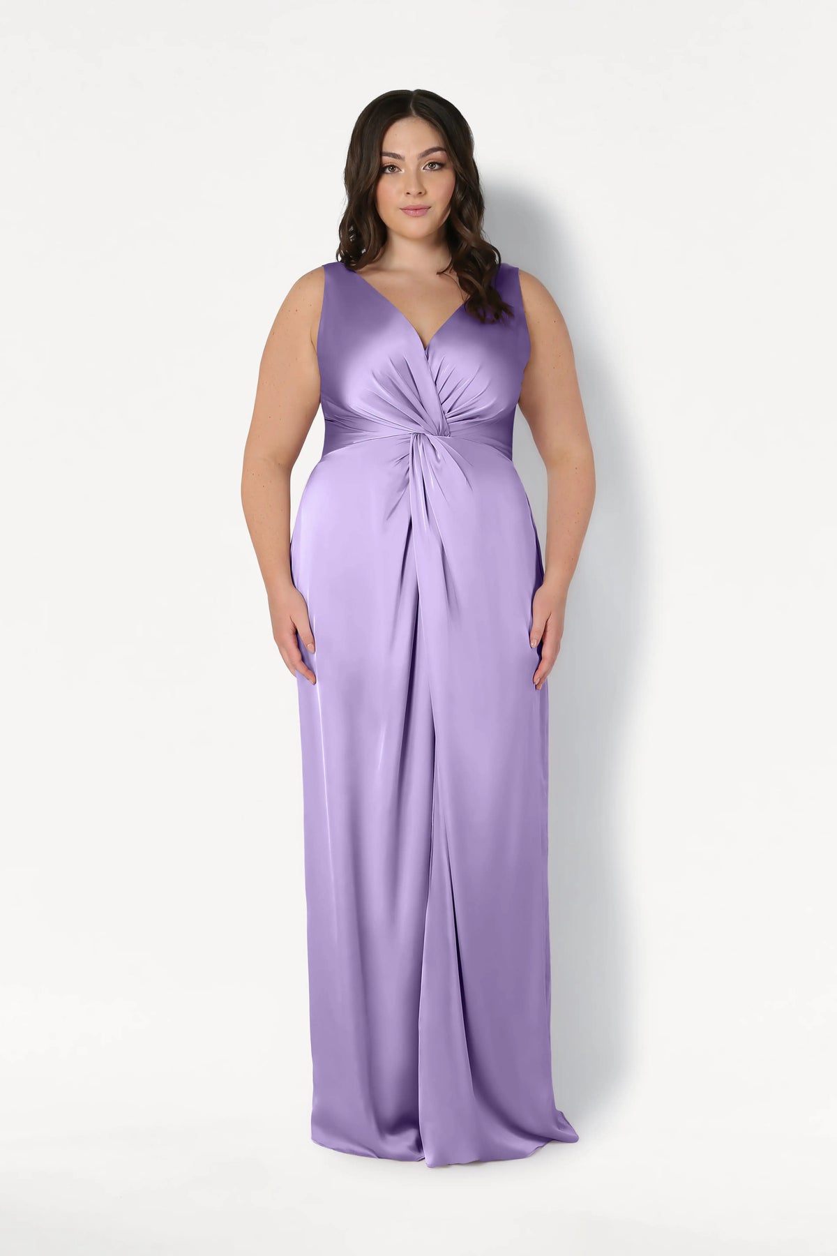 A woman with long, dark wavy hair wears the TH&TH Carmen Bridesmaid Dress in Violet Satin, a sleeveless floor-length gown with a V-neckline and twisted waist knot, standing against a plain white background.
