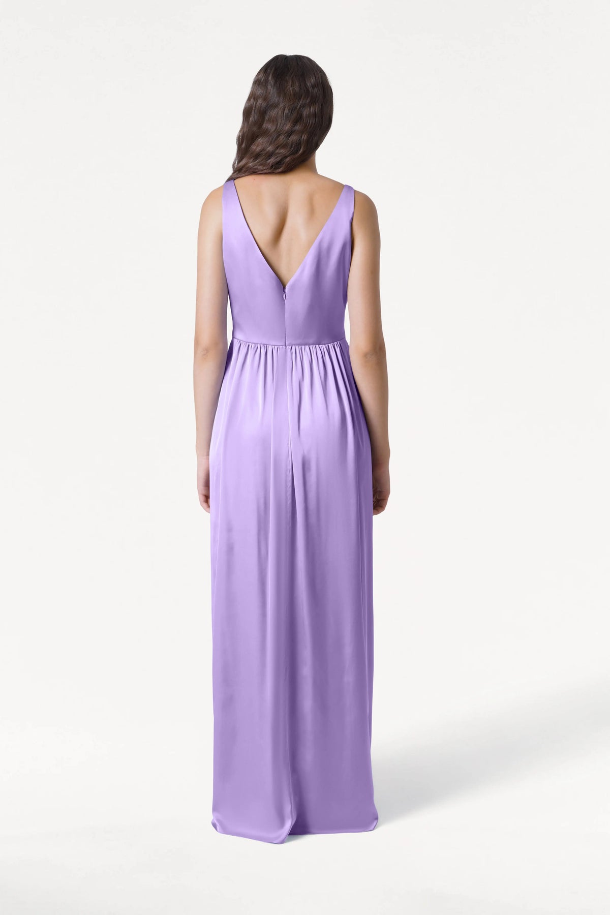 A woman with long wavy hair stands with her back to the camera, highlighting the sleeveless, floor-length Carmen Bridesmaid Dress in Violet Satin by TH&TH, featuring a deep V-back. The background is plain white.