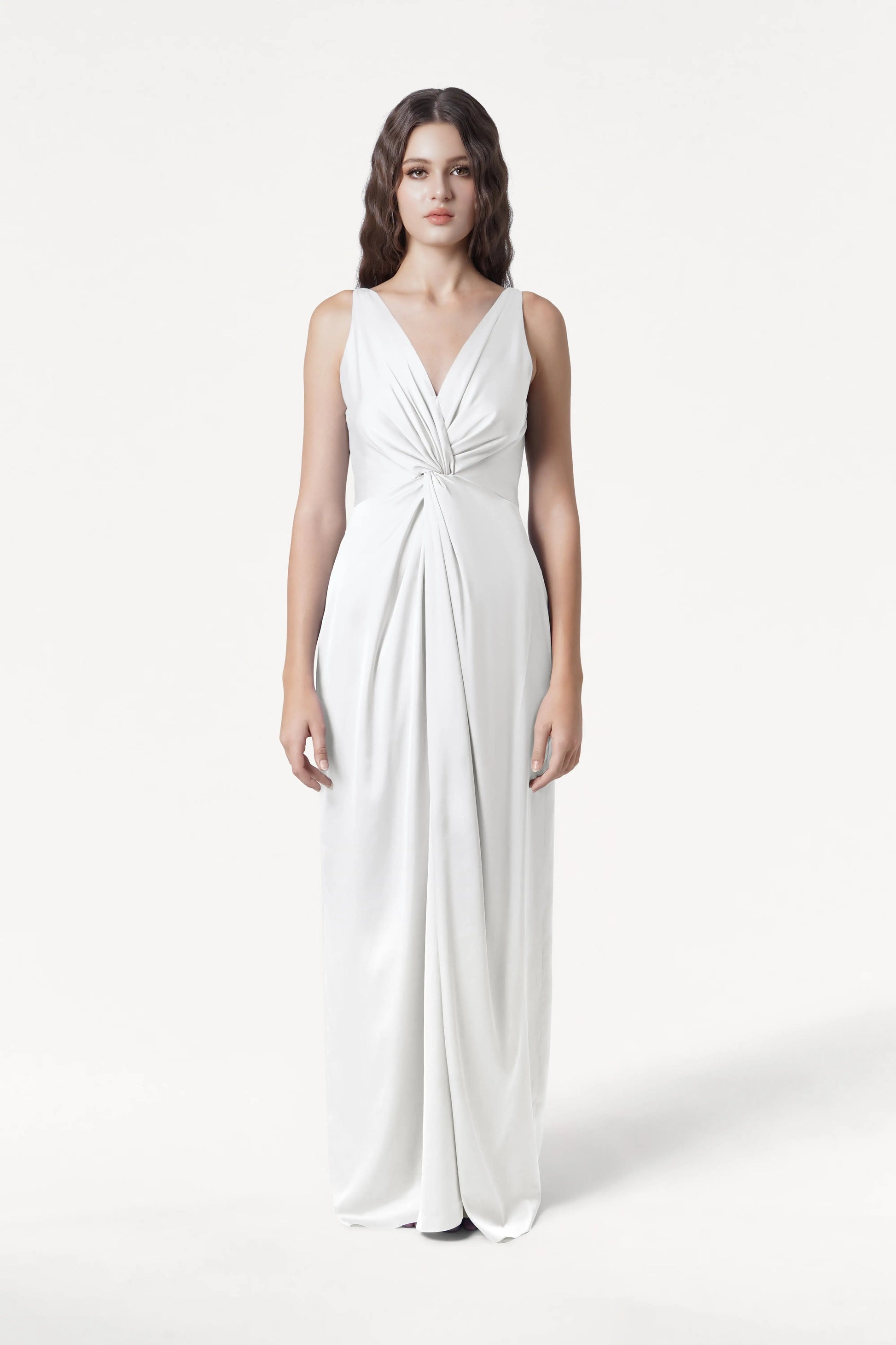 A woman with long wavy brown hair stands facing forward, wearing the TH&TH Carmen Dress in Ivory Satin—a sleeveless, floor-length gown with a V-neckline and twist-knot waist—set against a plain white background.